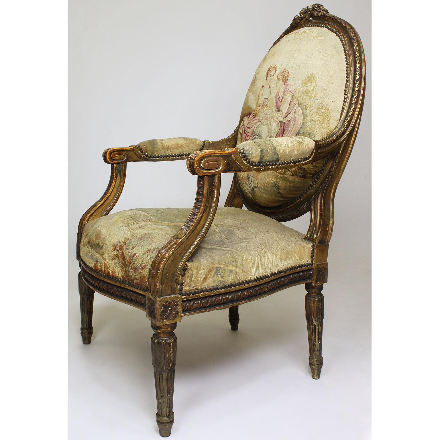 A set of four French 19th century Louis XVI style giltwood carved and Aubusson (worn) tapestry upholstered fauteuils a la reine armchairs. The banded oval back surmounted by a carved floral garland over leaf tip and scrolling acanthus carved padded