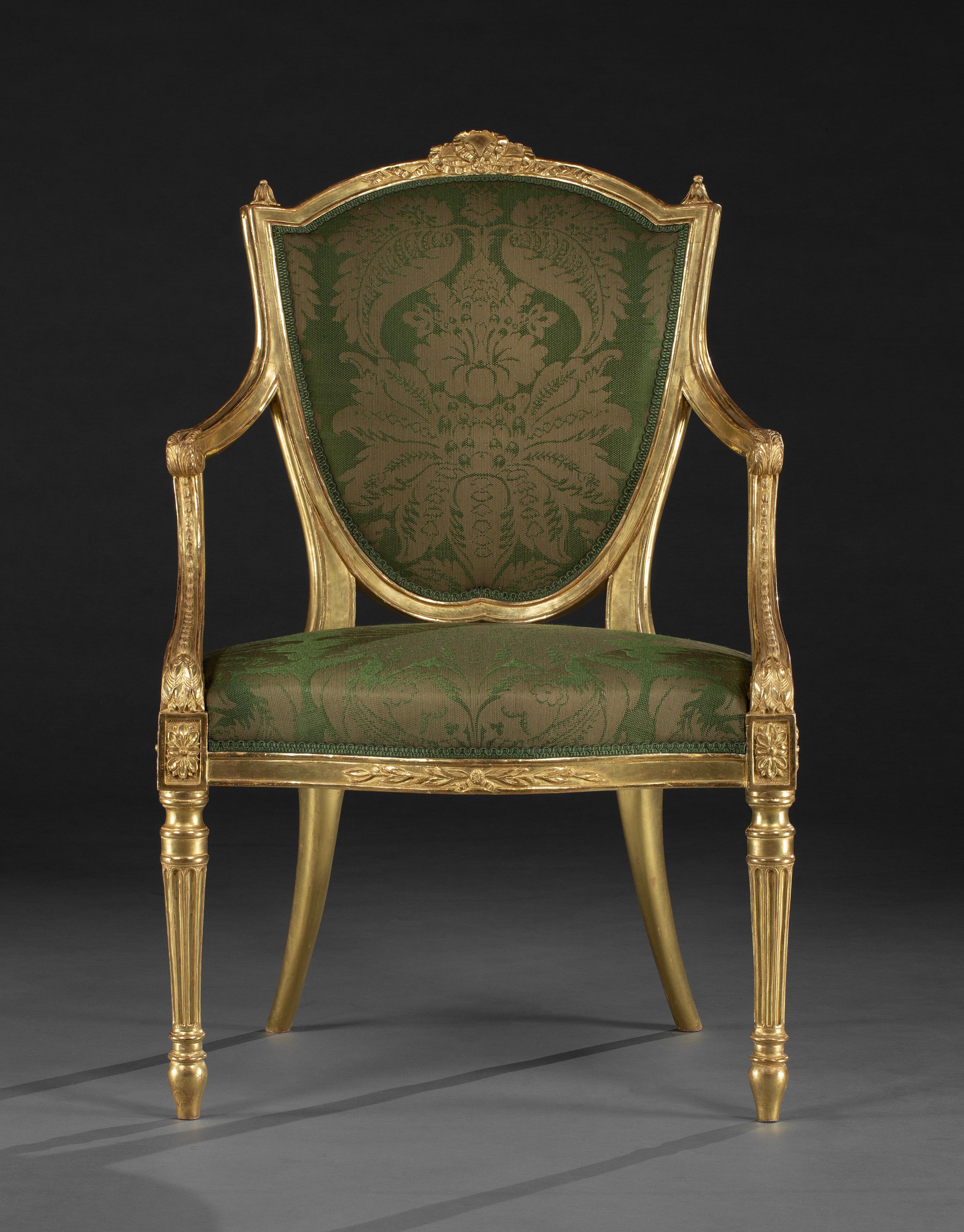 A fine set of four George III neo-classical giltwood armchairs. The shield shaped backs surmounted by a ribbon motif and finials to the uprights, the armrests terminating in acanthus scrolls, the padded backs and seats upholstered. The serpentine