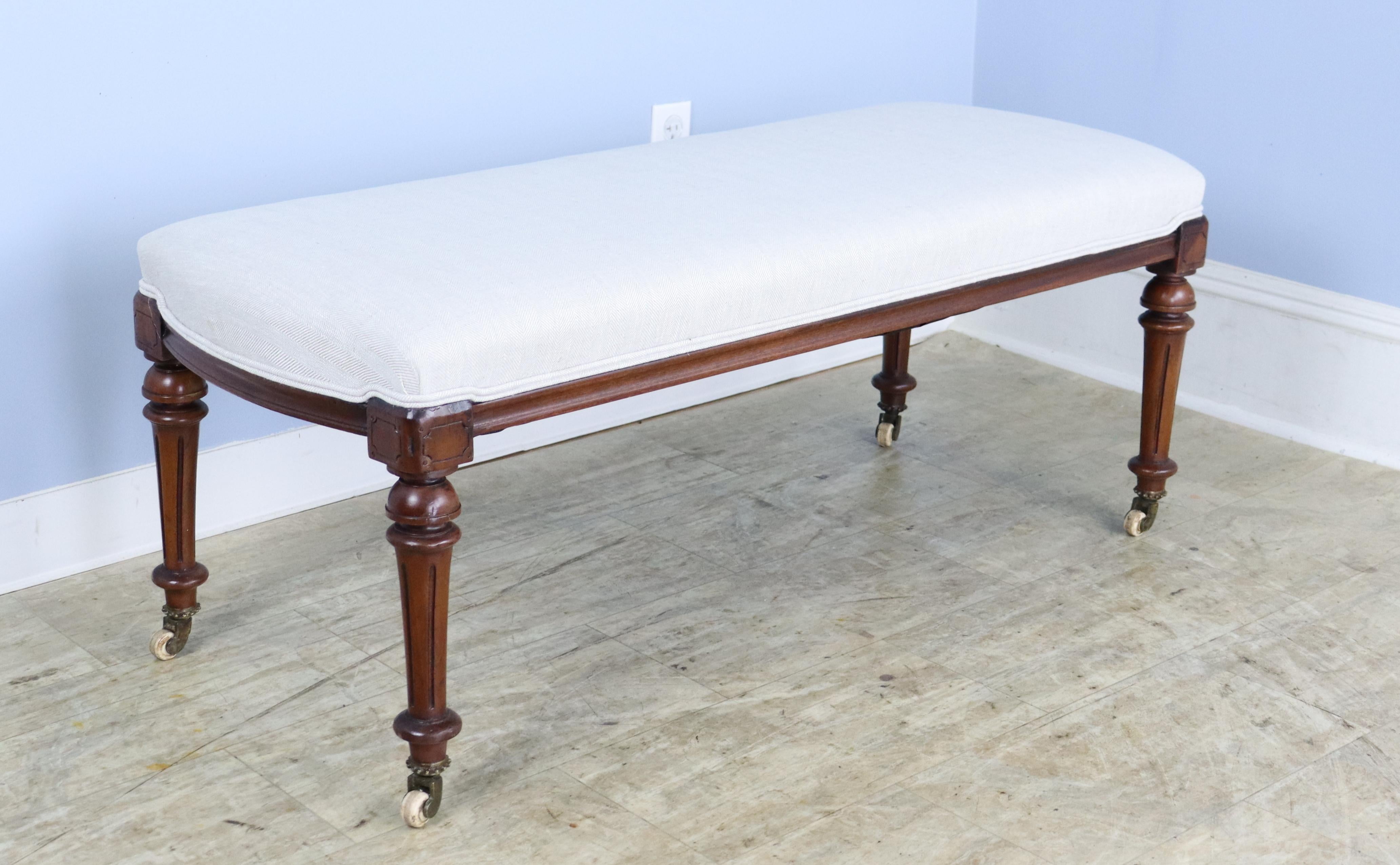 An elegant stool newly covered in French beige linen, with decorative piping at the edge of the seat. Constructed with mahogany legs, circa 1890 that are well-carved and sturdy.   Original castors.  Very good sitting-height, comfortable seating at