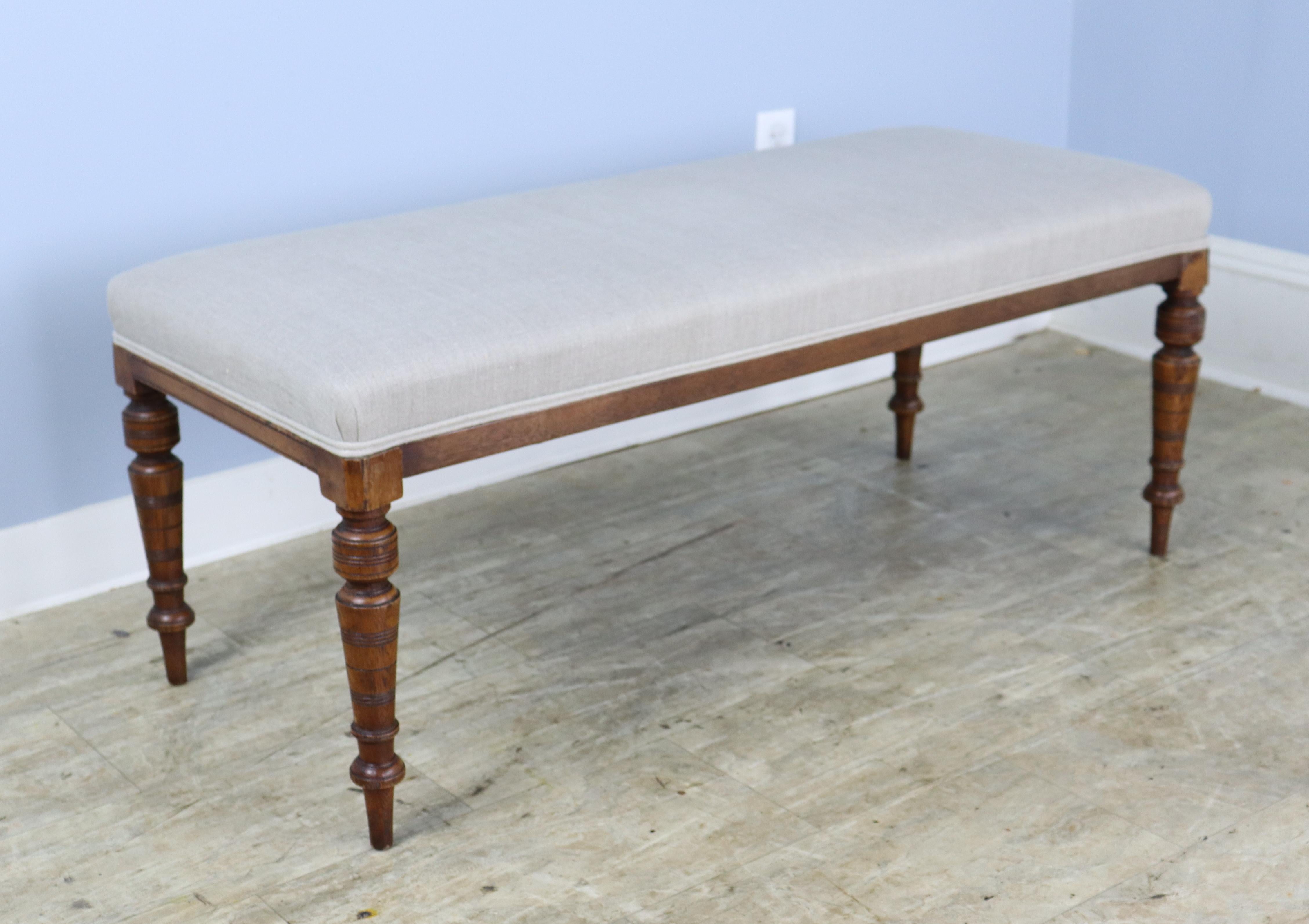 An elegant stool newly covered in French beige linen, with decorative piping at the edge of the seat. Constructed with mahogany legs, circa 1890 that are well-carved and sturdy. Very good sitting-height, comfortable seating at the end of the bed.