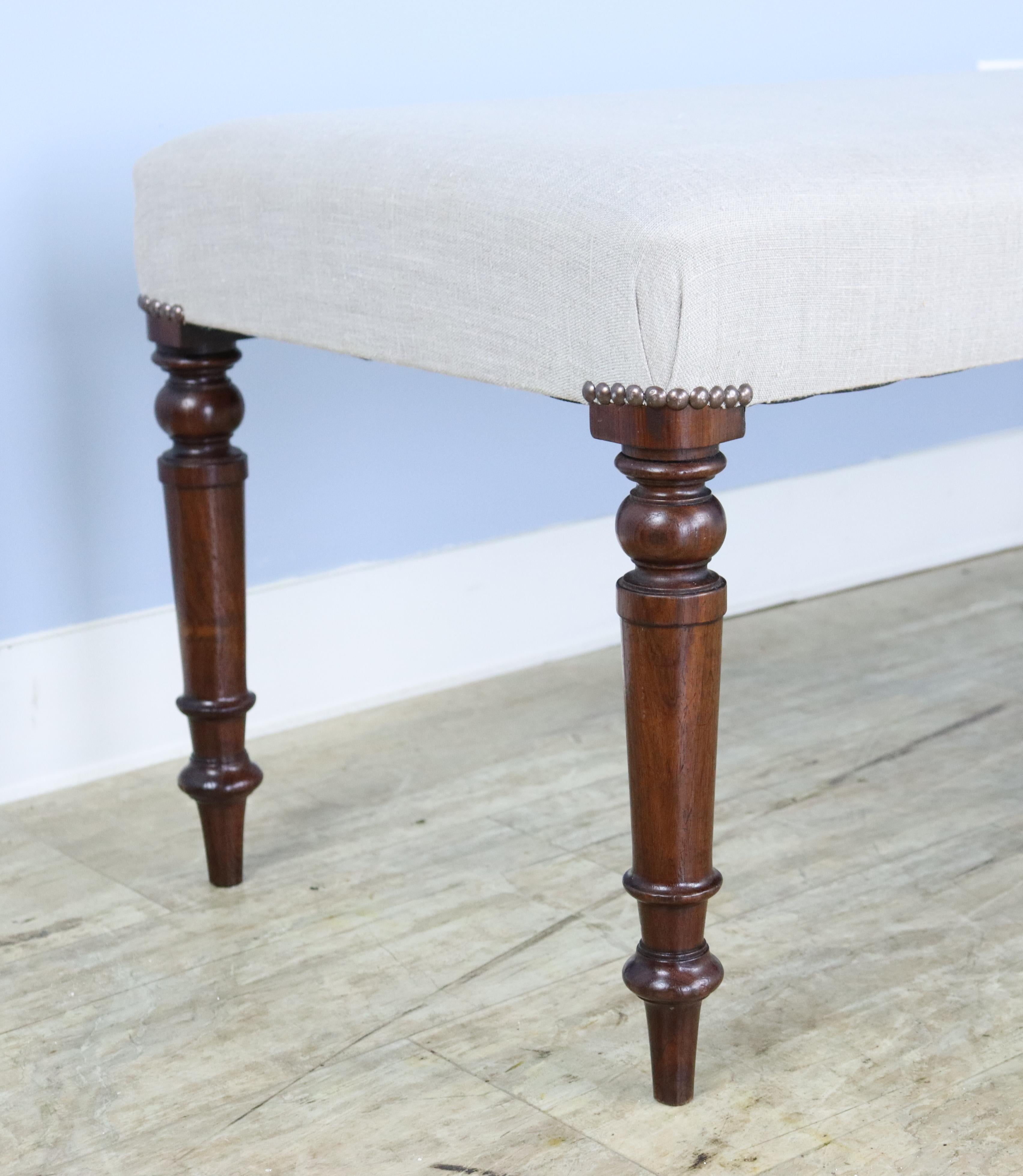 An elegant stool newly covered in French beige linen, with decorative nail heads at the edge of the seat. Constructed with mahogany legs, circa 1890 that are well-carved and sturdy. Very good sitting-height, comfortable seating at the end of the