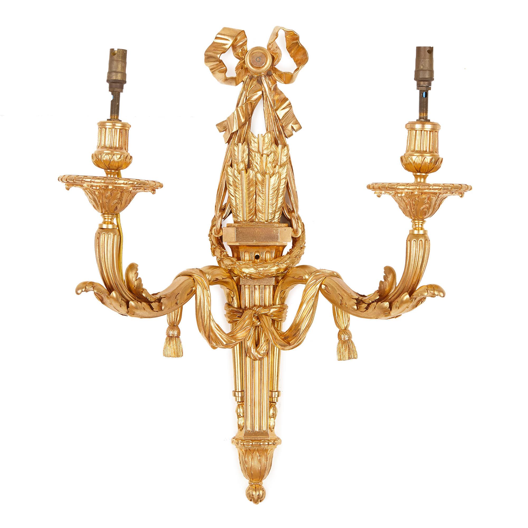 The gilt bronzes sconces are identically designed in an elegant neoclassical style. The stems of the sconces are shaped like arrow quivers, which are each mounted with two arms. These arms have foliate bases, which are draped with tasseled fabrics.