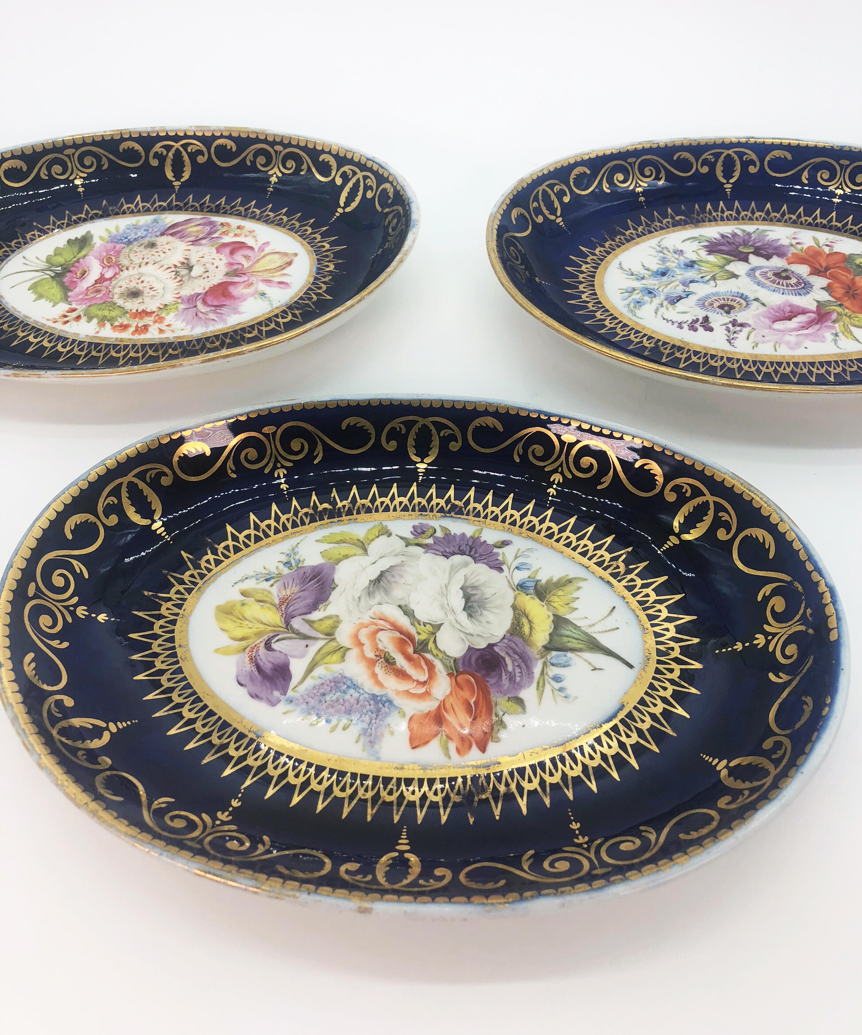 Set of four Coalport dishes consisting of three oval serving dishes and one scalloped dish all by Coalport with cobalt borders and gilded bands of interlocking scrolls and finials with a gold dentil border. Hand painted panels of floral bouquets in