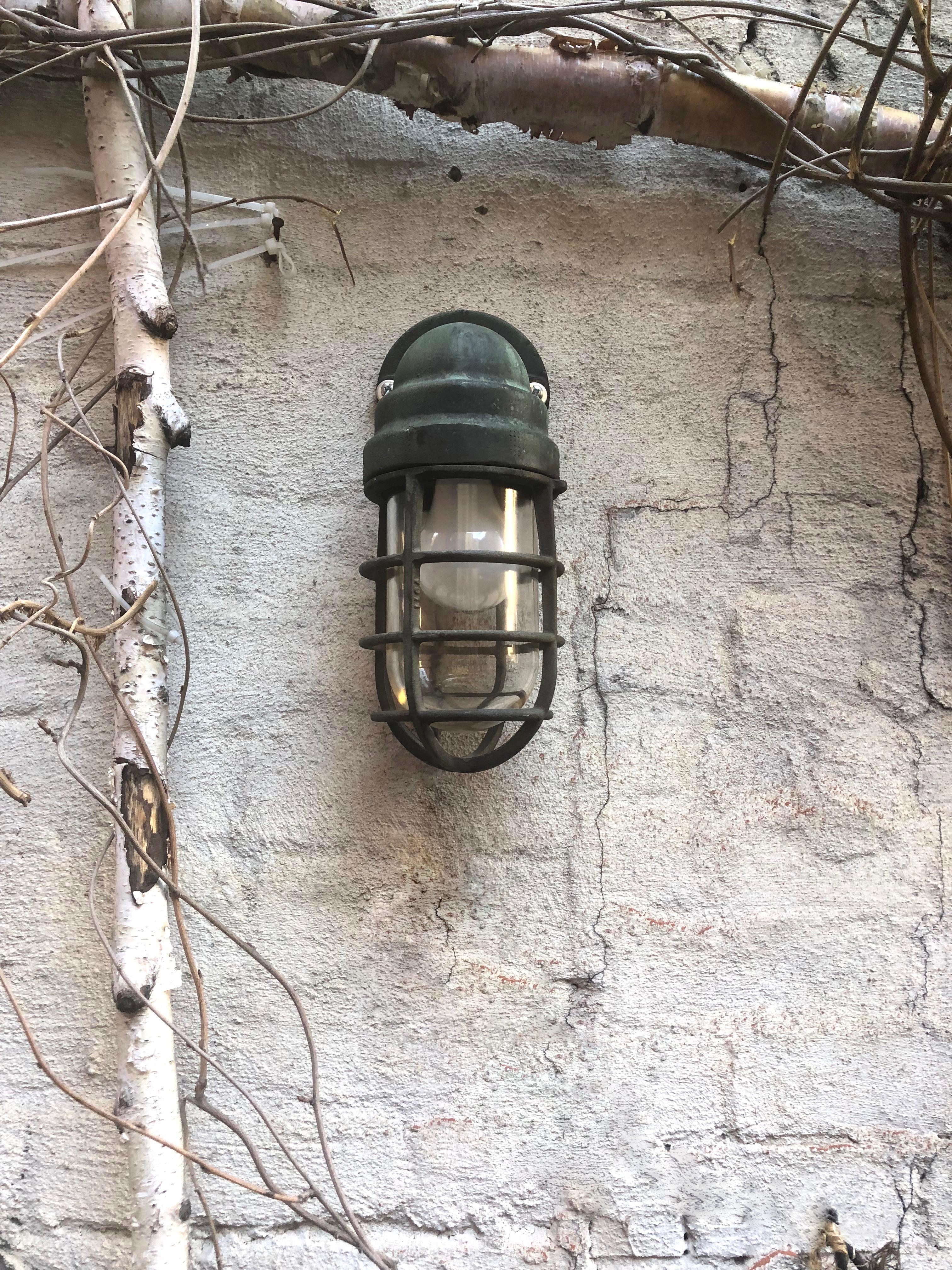 Four Crouse-Hinds industrial sconces in solid brass with its original, green patina and a shatter-proof clear glass shade enclosed within the metal frame.