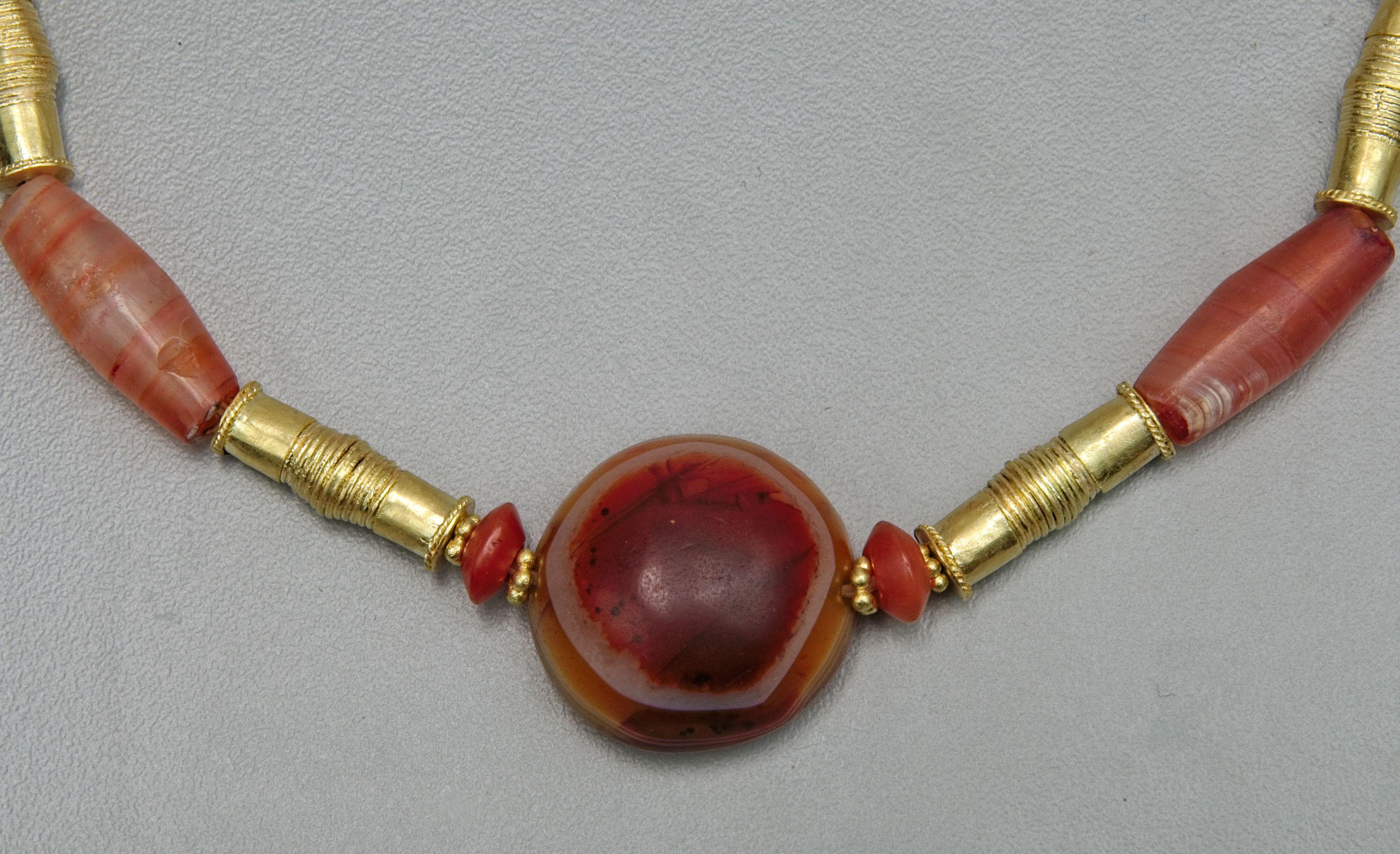 A necklace of six long carnelian tube beads, a round tabular carnelian “eye” bead, and four carnelian truncated bi cone beads. The carnelian tube beads alternate with six 20k gold beads. The small truncated bi cone carnelian beads are each faced