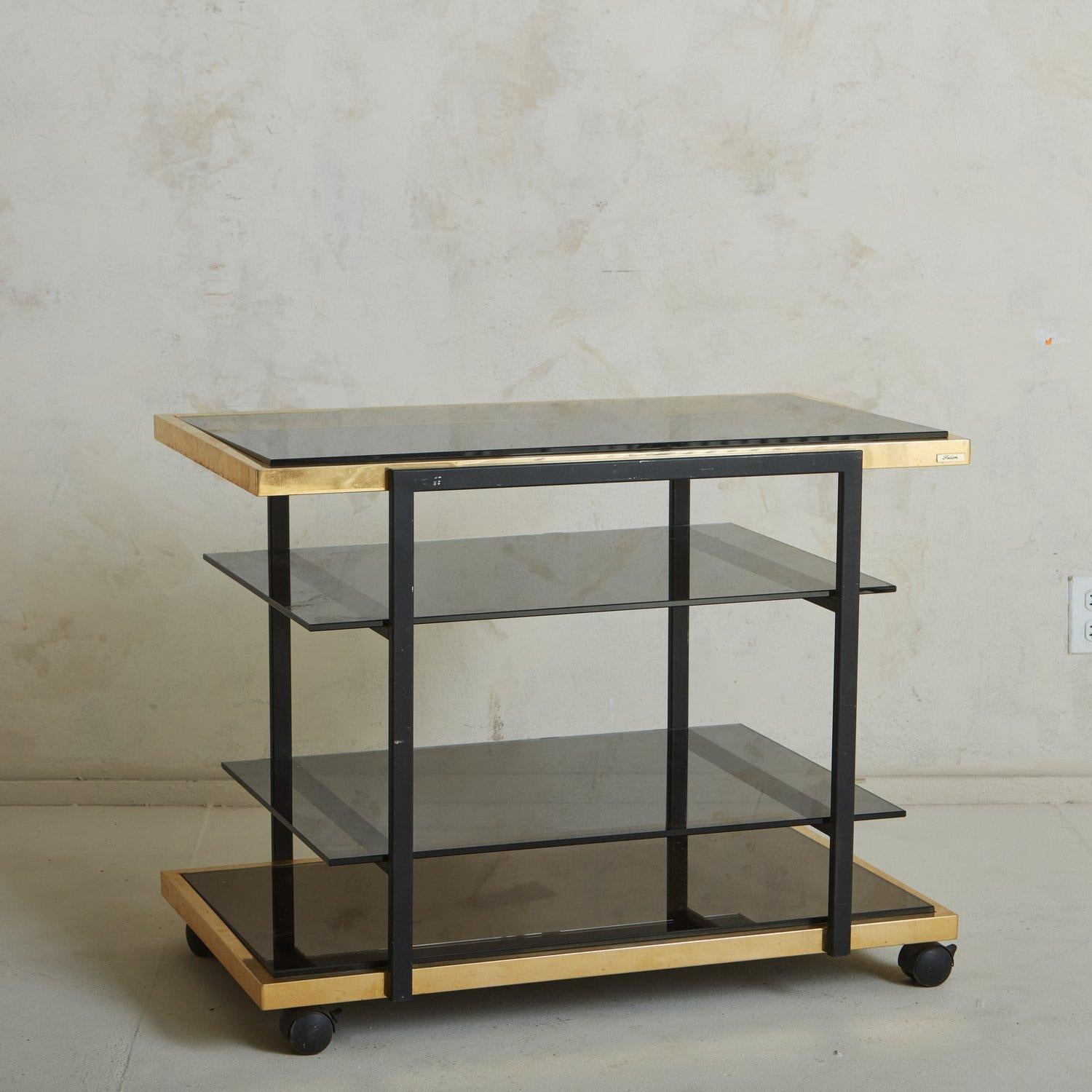 A vintage Belgian bar cart by Fedam featuring a brass and black metal frame with four smoked glass shelves. This cart is on casters for easy mobility and offers significant storage space, perfect for styling barware, bottles and books. Retains