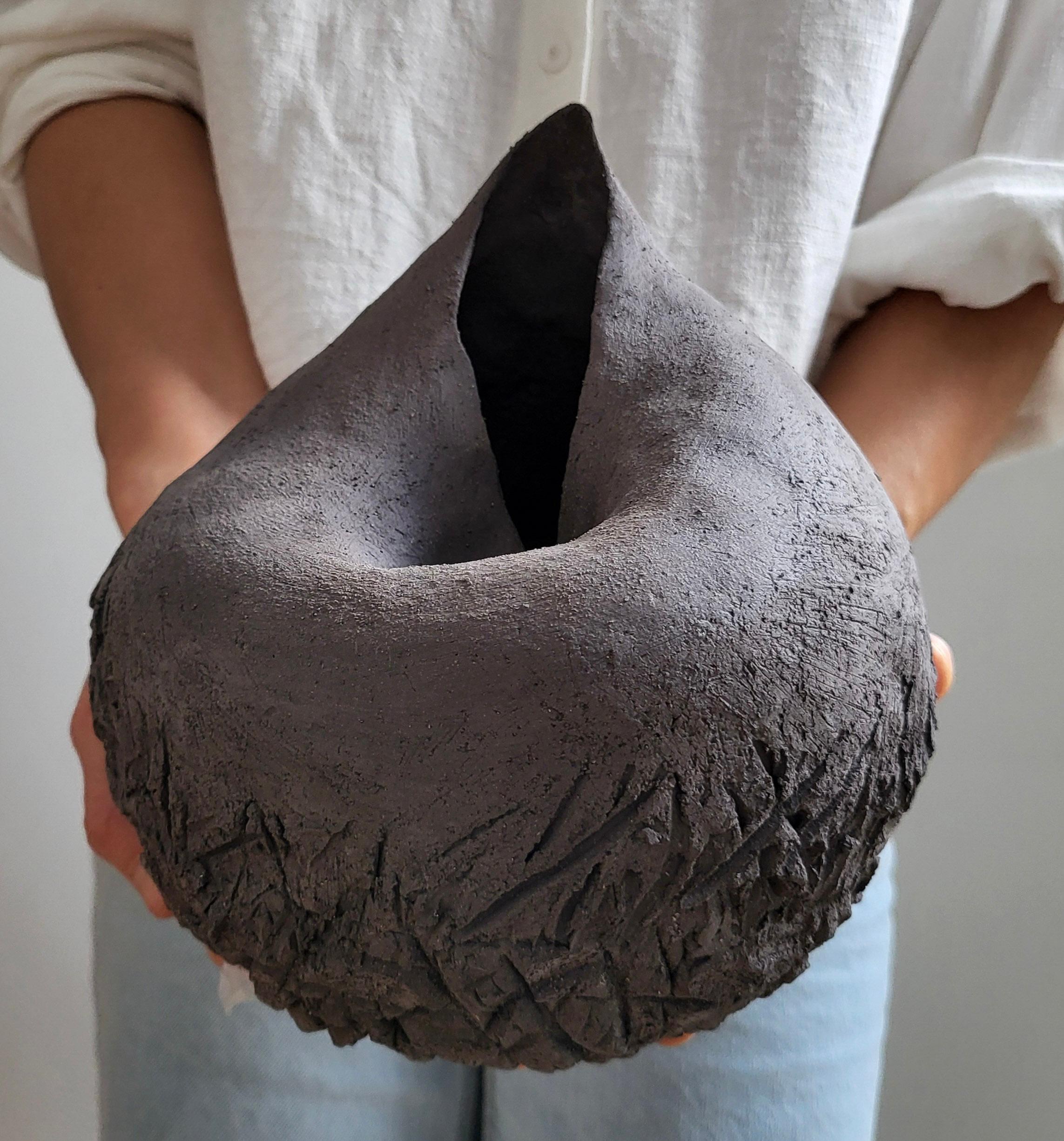 Fragment of the Earth is a hand-built ceramic sculpture by French artist Audrey Vo Van, shaped in black chamotte stoneware. The piece combines smooth, rounded contours with a deeply textured base, evoking the layered memory of geological
