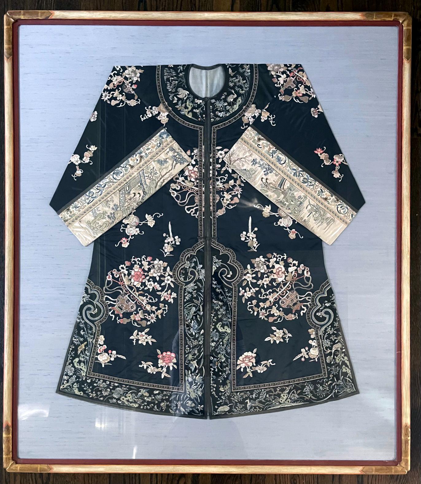 A woman's coat made of black silk with wide sleeves from Chinese late Qing Dynasty (mid to late 19th century), mounted and presented on blue linen board and framed as a stunning piece of textile art. The coat features white silk bands on wide