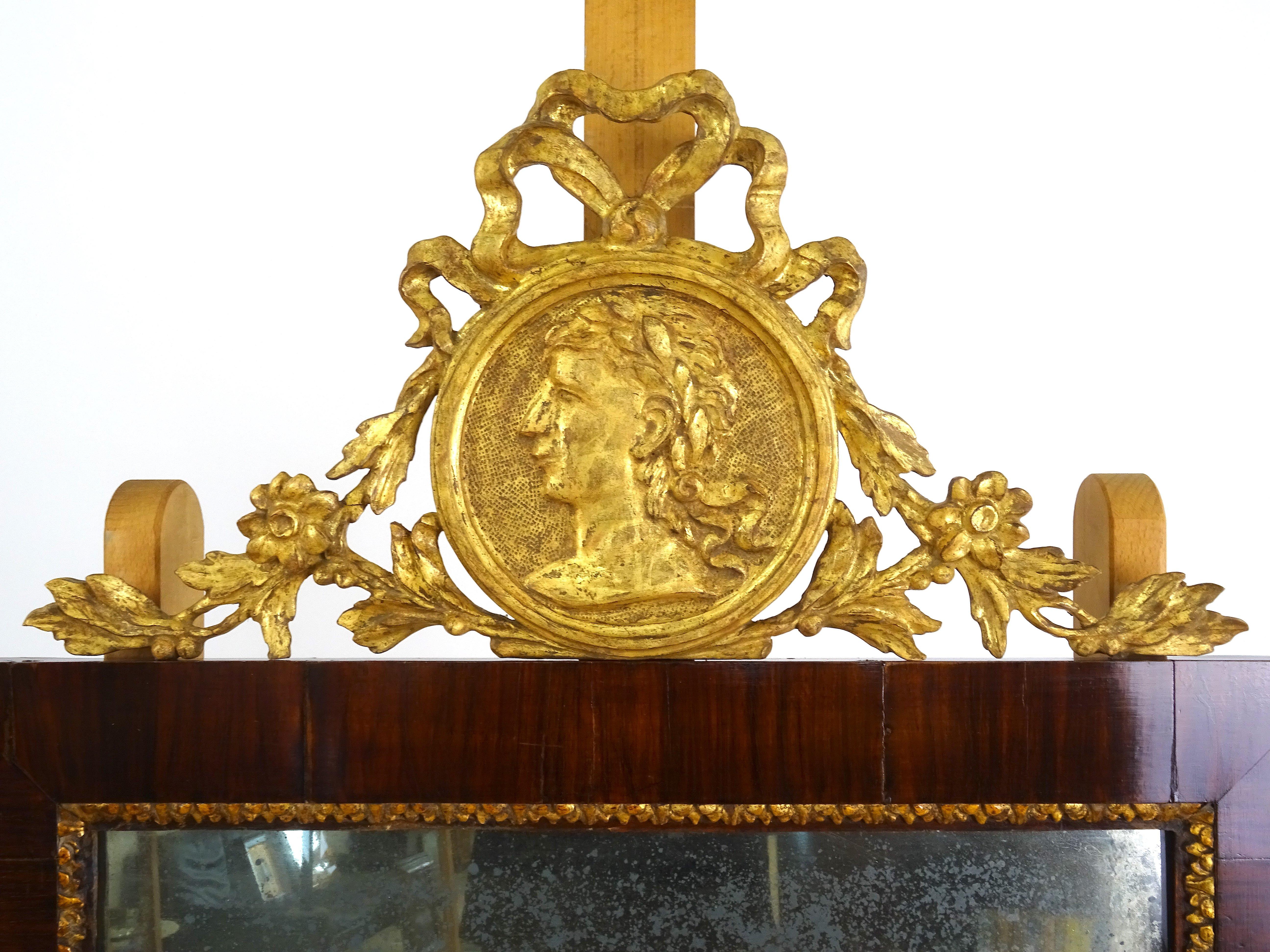 The piece in question is an elegant frame with a mirror, with a carved and gilded wooden molding in the upper part composed of a floral decoration and a central medallion depicting a face in profile.

The wooden frame and mahogany veneer, is of