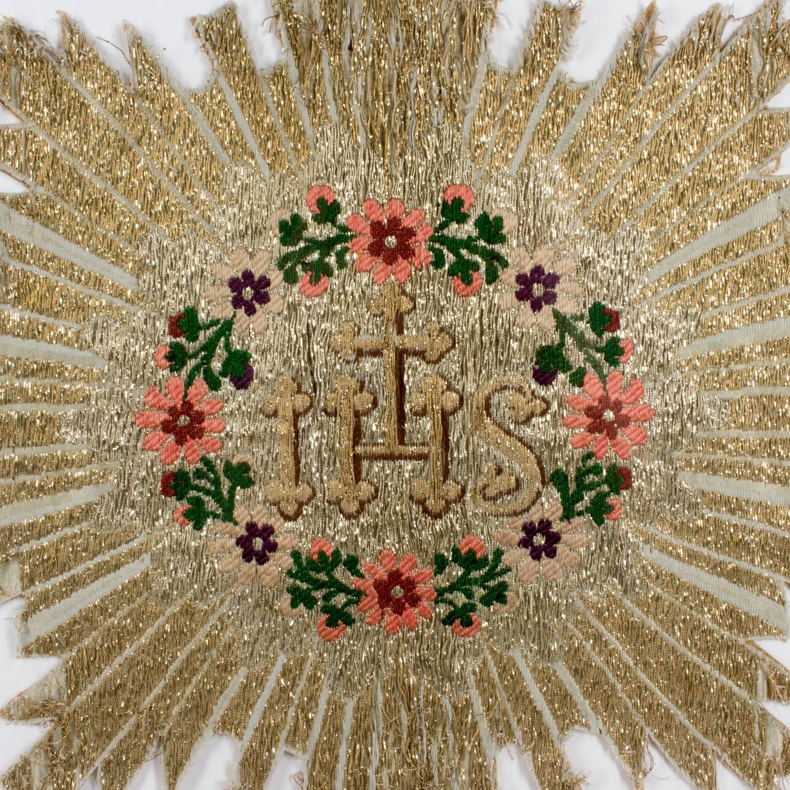 This beautifully made antique (late 1800s) textile fragment shows a metallic starburst shape with colorful embroidery at the centre, including flowers and the letters IHS, commonly known as a Christogram, which represents Christ. This piece was