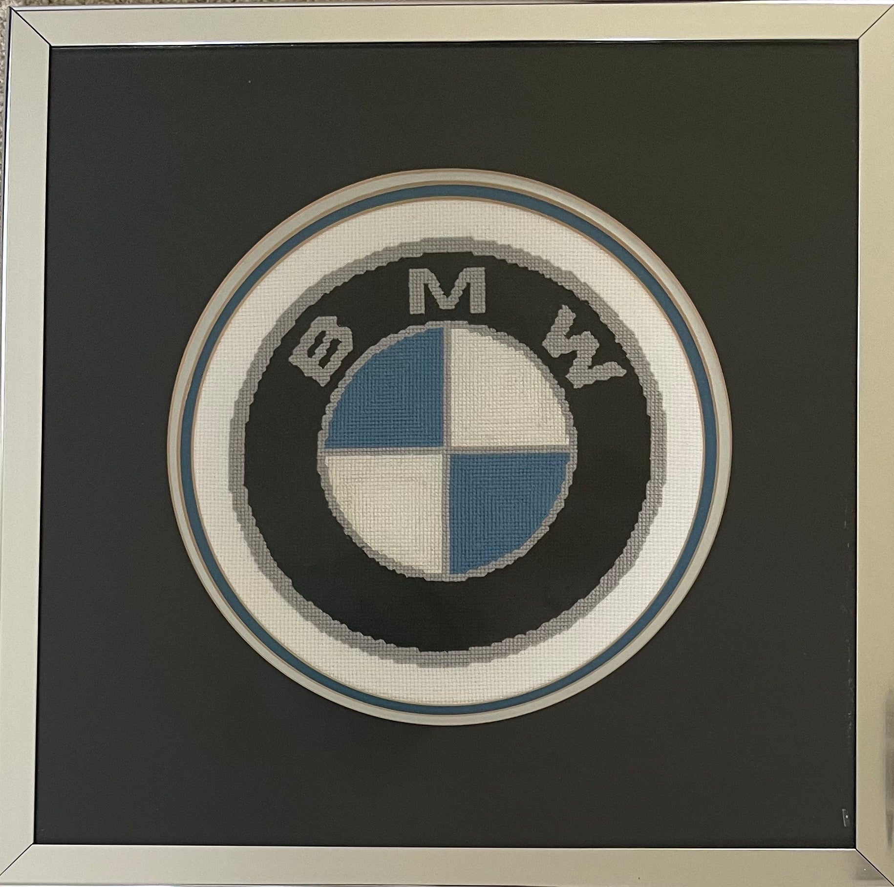 A very well done BMW embroidered logo housed in a silver frame with glass and hanging wire to back. The piece is in very good condition and measures 17