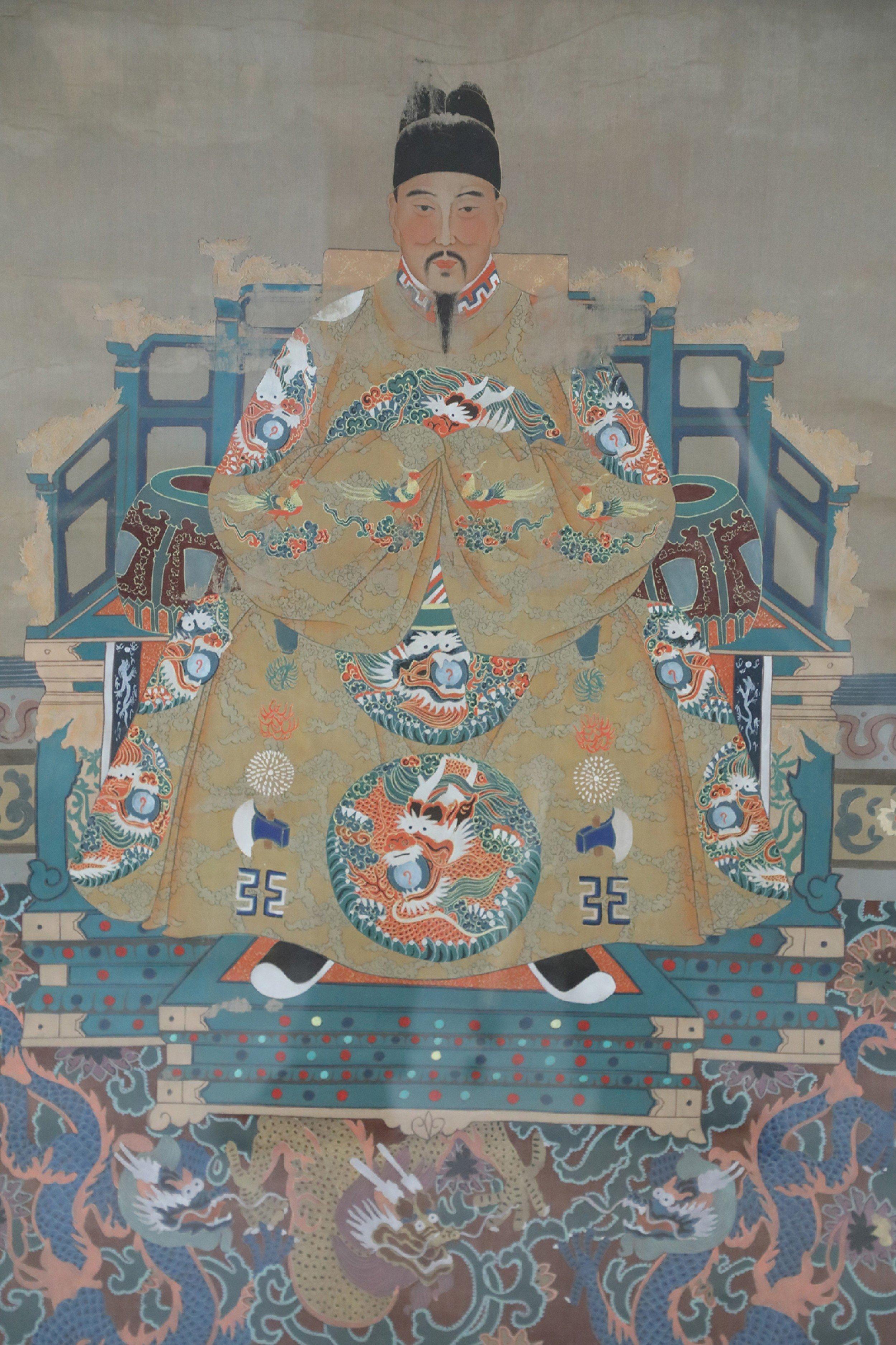 Chinese ancestor portrait depicting a man in flowing golden robes decorated with medalions featuring dragons, seated above a floor featuring additional dragon designs in a heavy rectangular dark wooden frame.
  