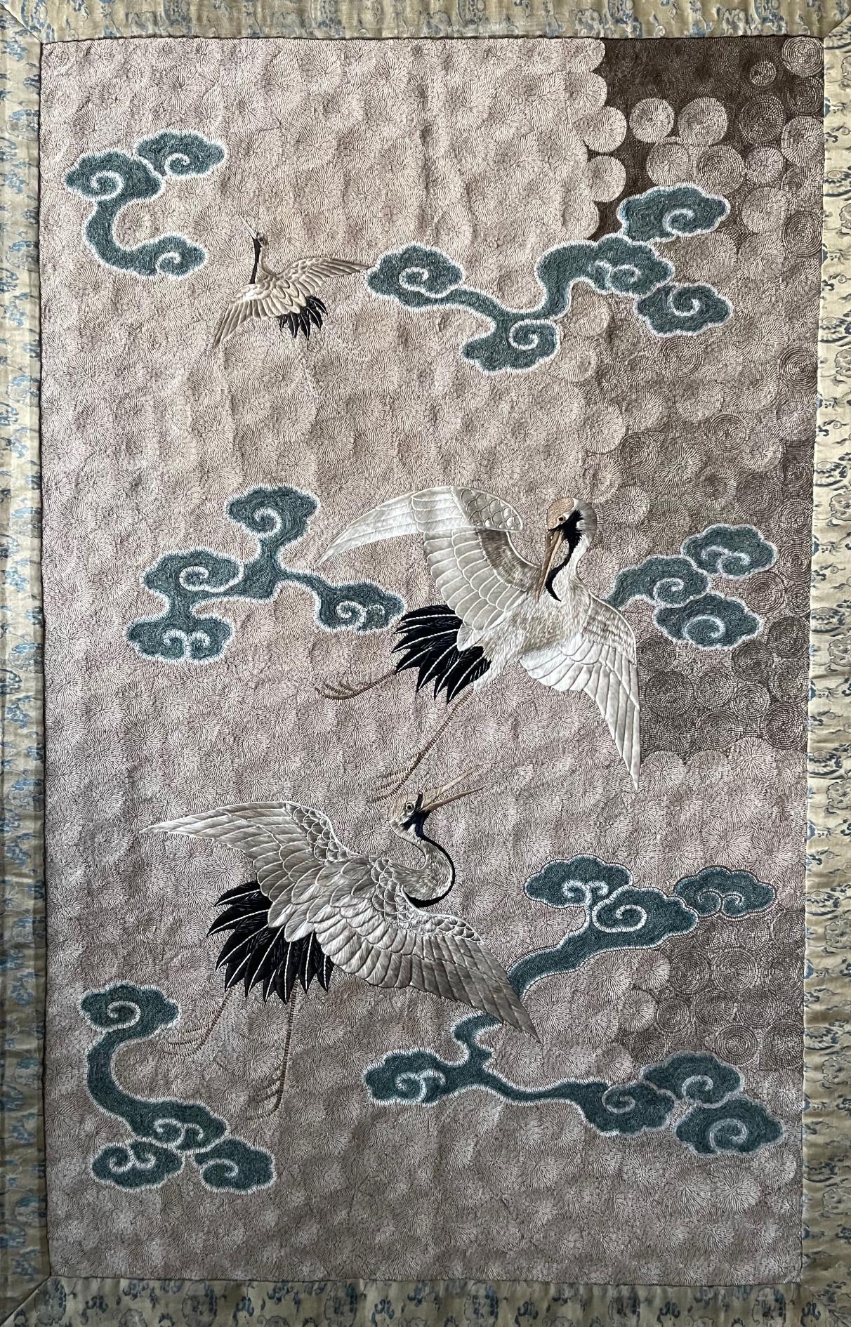 A Japanese embroidered textile panel presented in an acrylic shadow box frame, circa end of 19th to 1910s Meiji Period. The textile panel depicts three flying cranes among auspicious clouds, one of the most popular subject in Japanese art. Crane is