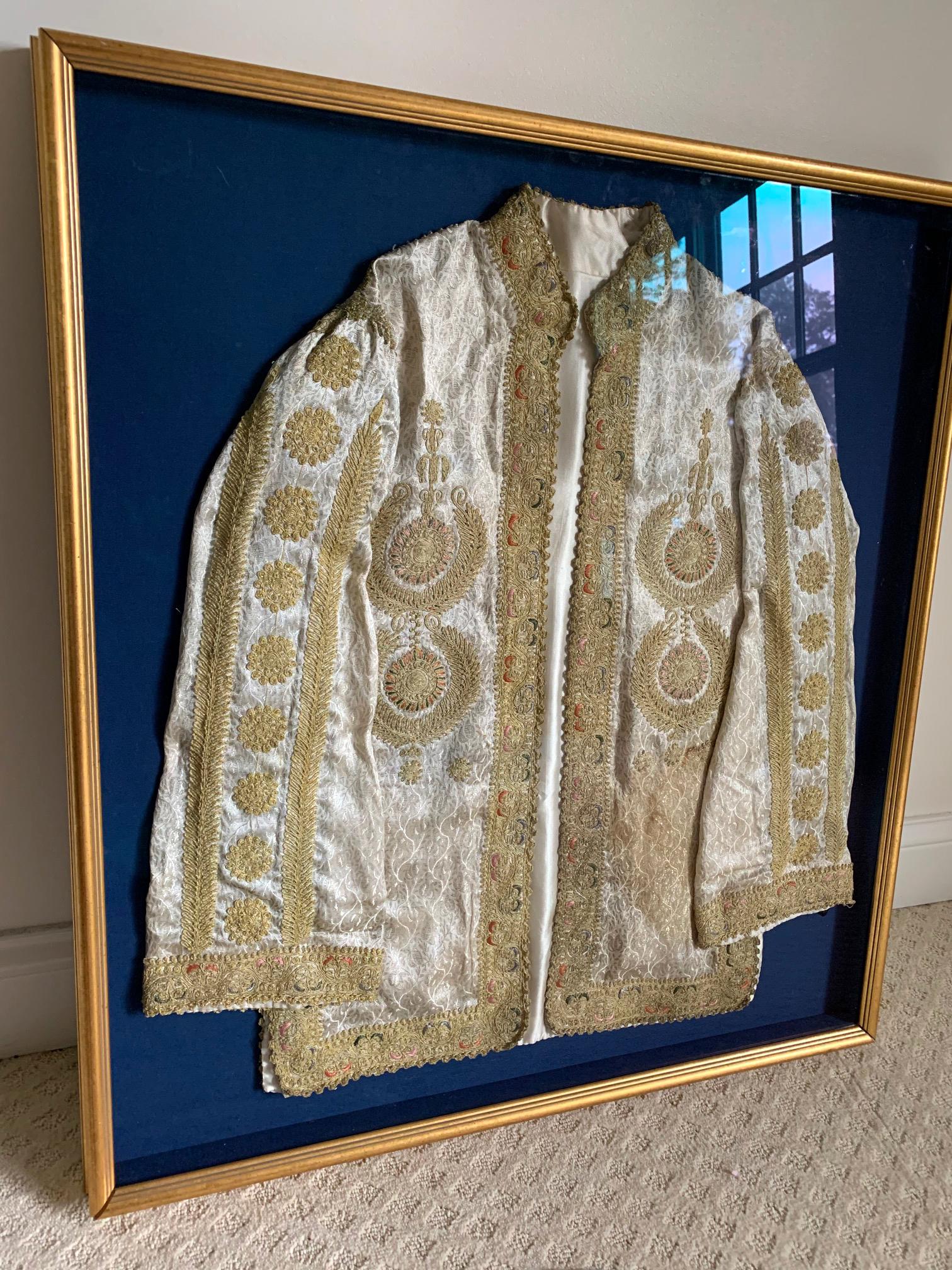 An lavishly decorated Turkish coat likely from the Ottoman Empire period circa 19th century, presented in a shadow box gilt frame with dark blue fabric lining. The jacket was made from a silver damask patterned brocade with elaborate embroidery with