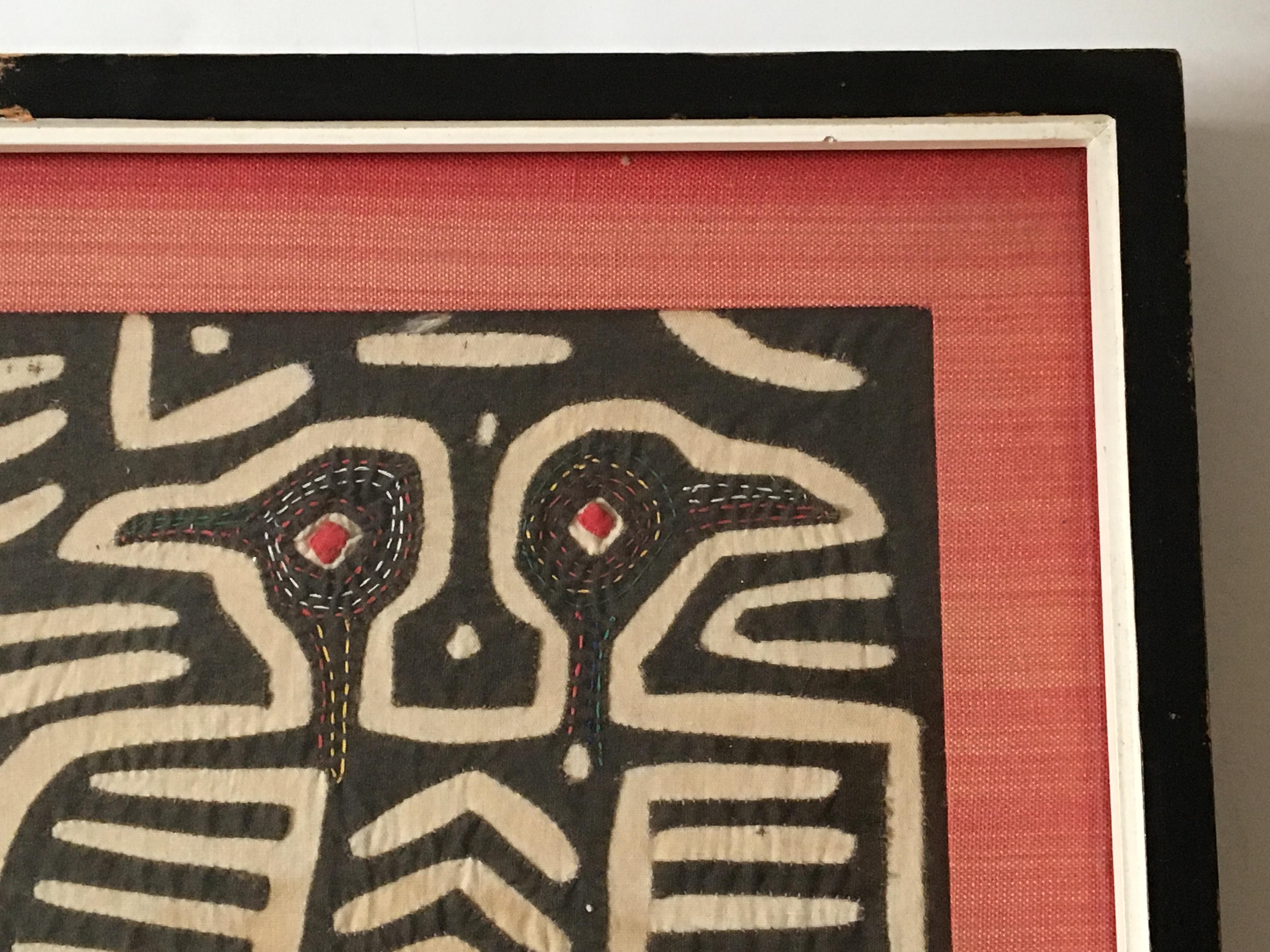 A petite framed kuna mola, the traditional Panamanian costume, which depicts the most intriguing symbolism of a pair of two-headed birds with red eyes. There is even more detailing in the form of multicolored stitching all around their head and neck