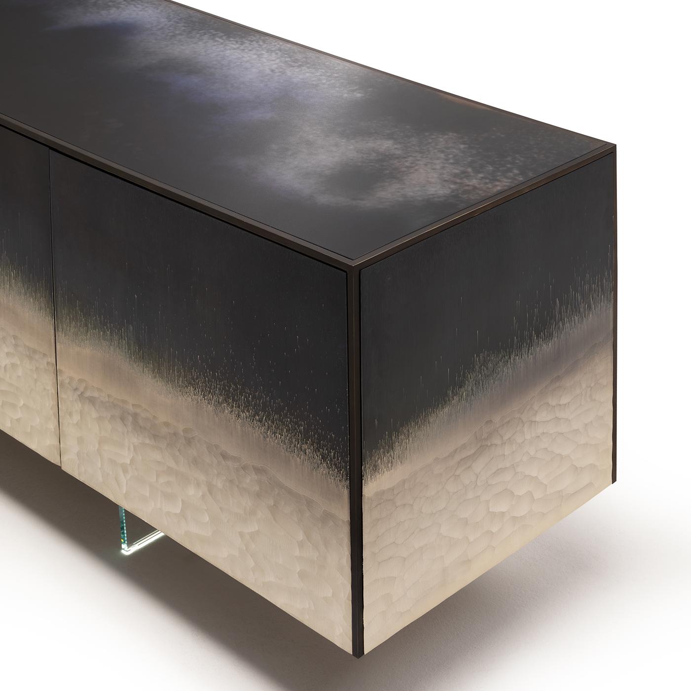 Struktur aus Esche furniertem Sperrholz mit Metallic-Lackierung in Titan. Massives Metallprofil aus dunkel brüniertem Messing. Türen, Seiten und Deckel mit Stromboli-Finish verziert. Vier Türen, innen ein extra klarer Glasboden mit abgeschrägter