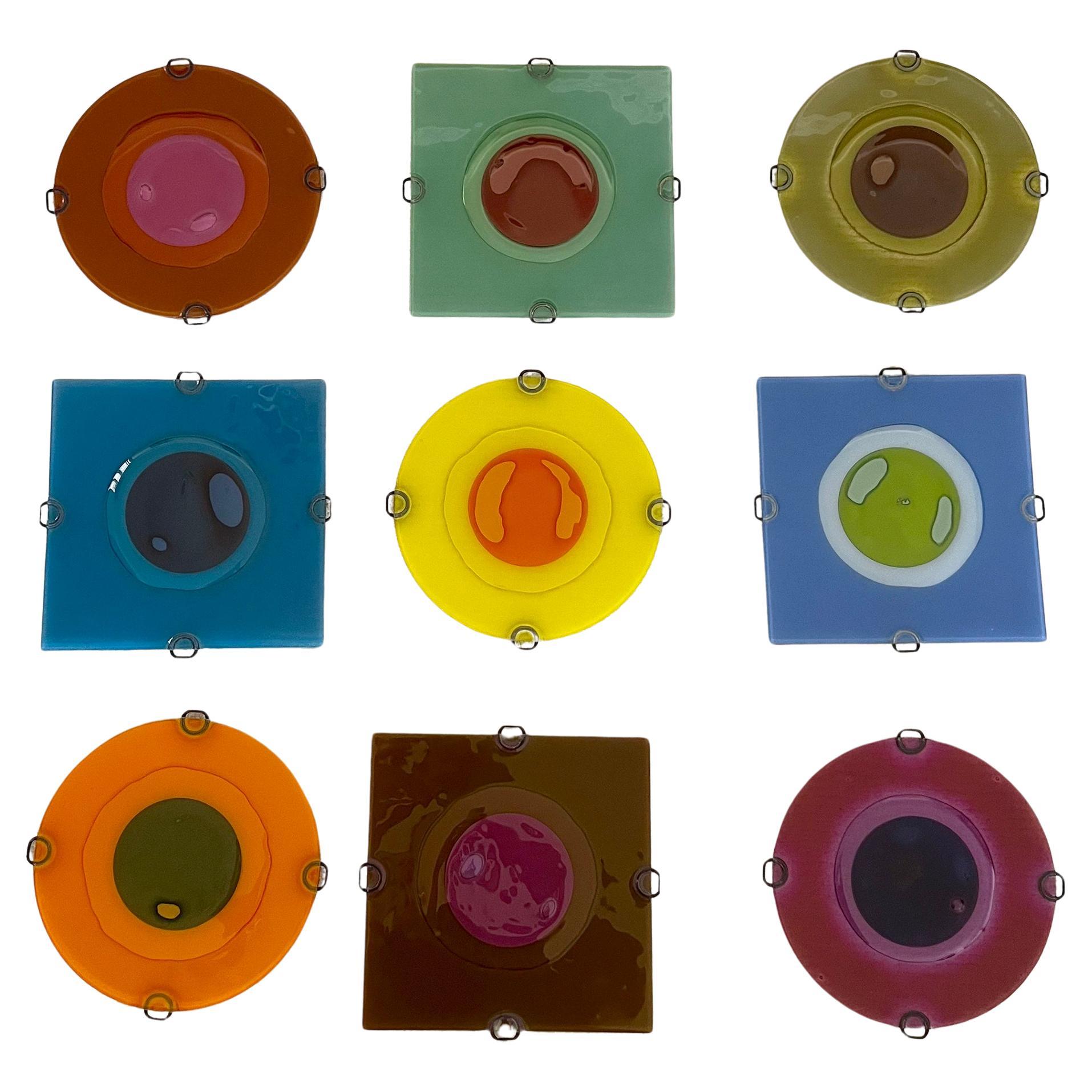 Fifteen fused 9″ circles of richly-colored handcrafted glass in square and circular shapes, created by Frances and Michael Higgins of Riverside, Illinois. These date from the mid 90's and do include the metal springs which attach the pieces