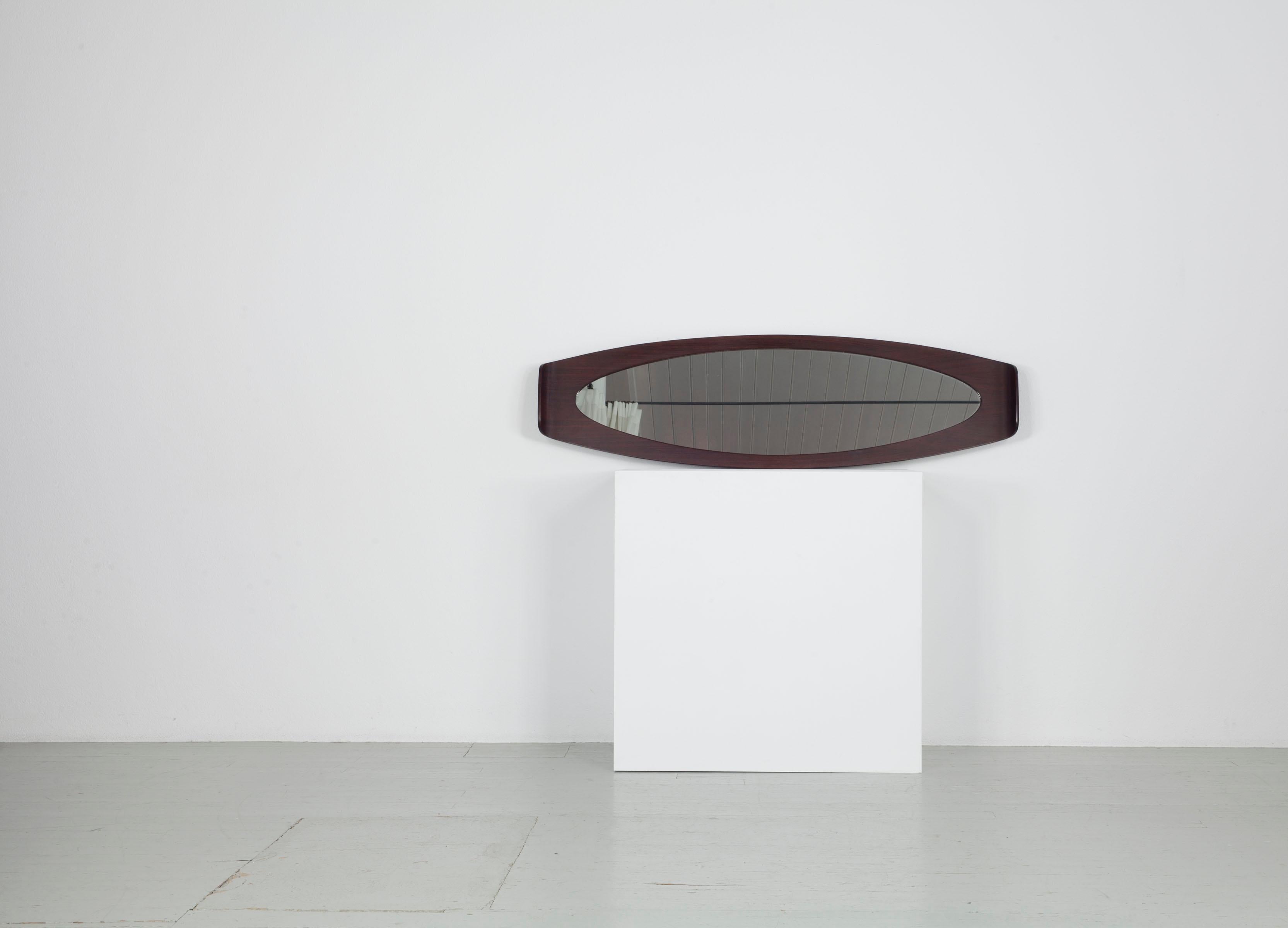 Dieser italienische, langgestreckte, ovale Spiegel stammt aus den 60er Jahren. Es wurde von Franco Campo und Carlo Graffi entworfen.  Der Spiegel hat eine ganz besondere Form, die ovale Länge des dunkel gebeizten Palisanderholzes ist an beiden Enden