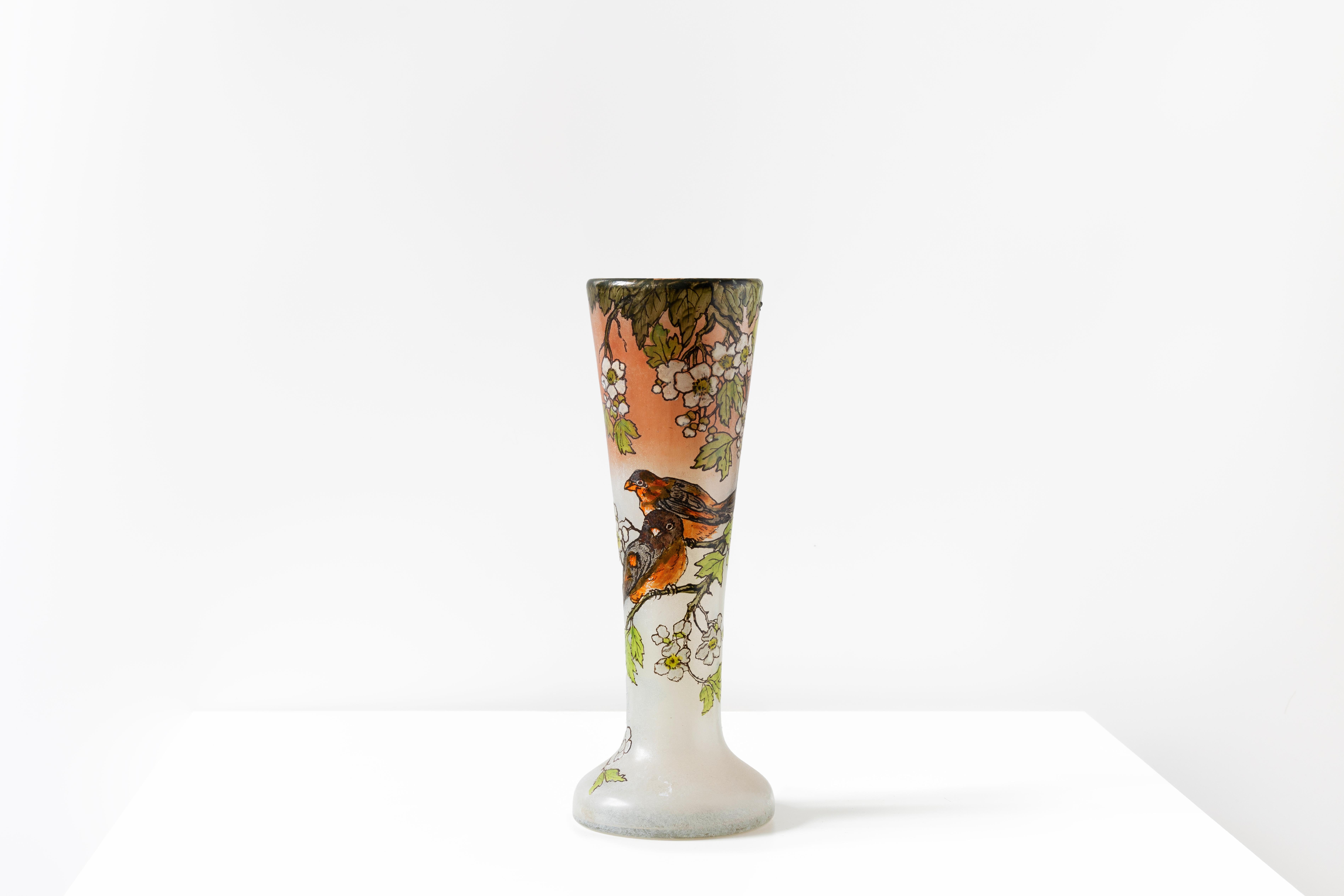 Questo splendido vaso Art Nouveau di François-Théodore Legras, realizzato in vetro bianco e rosato, è un pezzo elegante che presenta un collo graziosamente svasato e una base circolare, ornata da una scena finemente smaltata di due uccelli