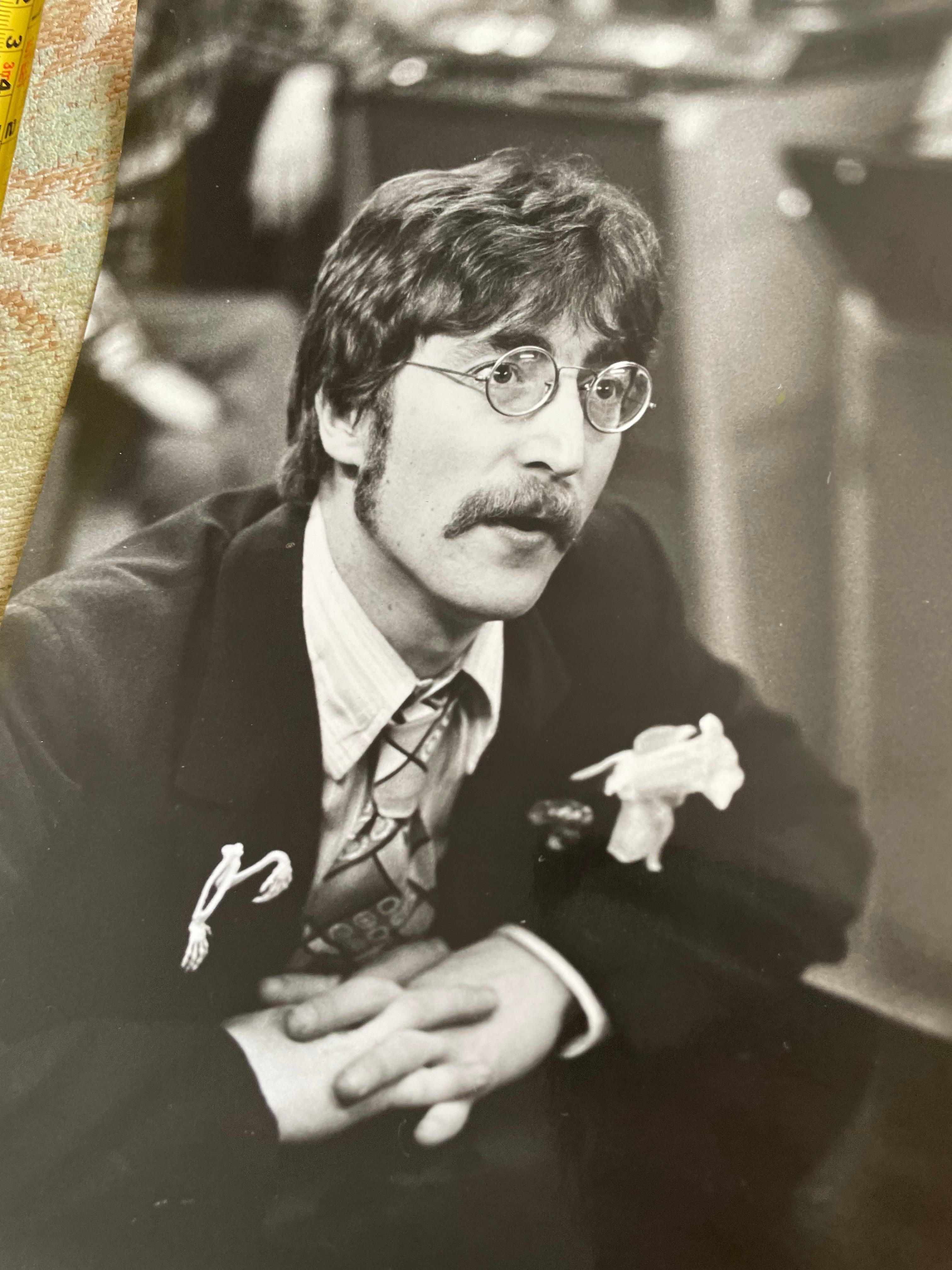 John Lennon negli Studio Abbey Rd di Londra durante la registrazione di Sgt Pepper's Lonely Hearst Club band, 1967. Seduto appoggiato alla scrivania della console, indossa un fiore sul bavero sinistro, una spilla scheletrica sul bavero destro e i