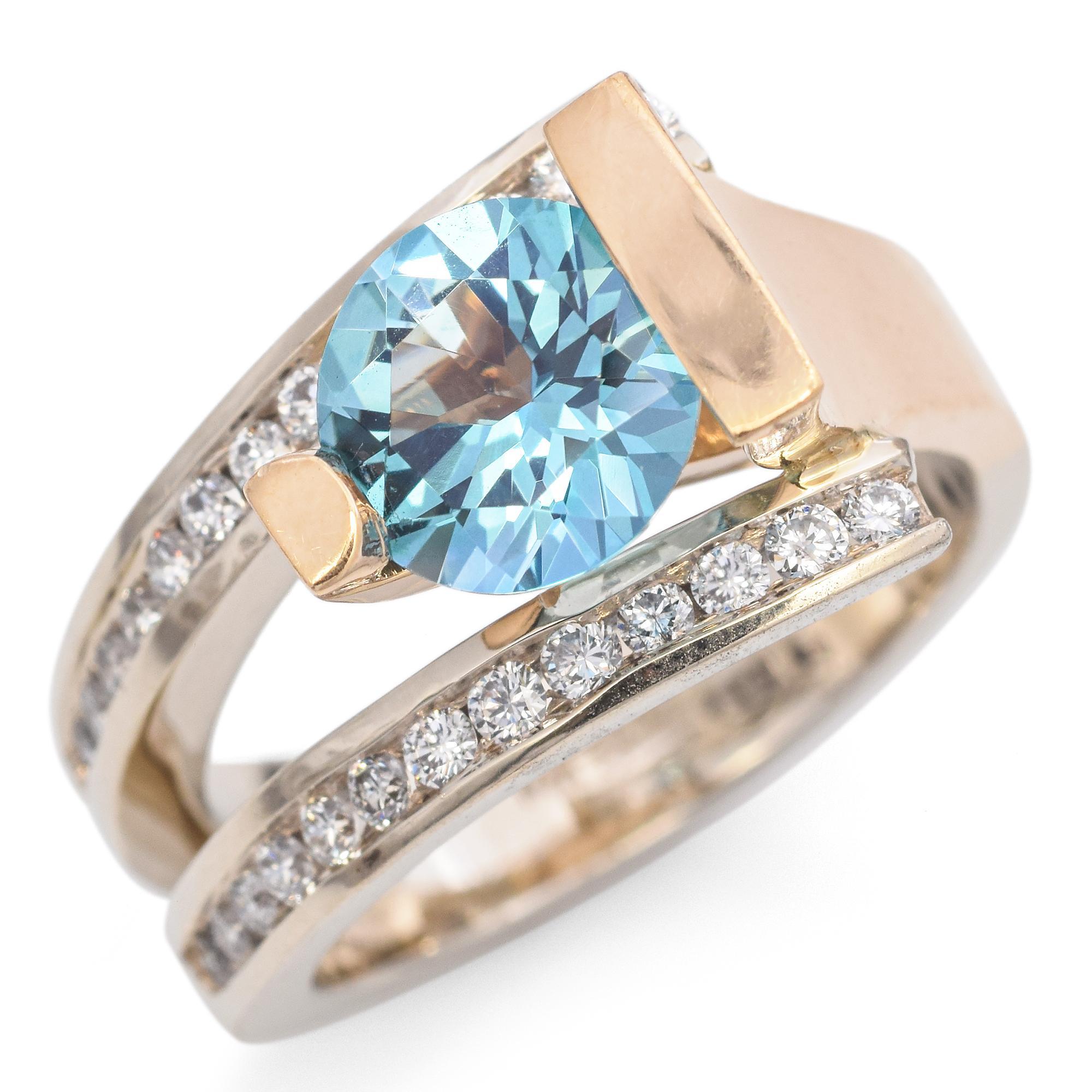 Ein auffälliger, moderner Ring aus 14 Karat Gelbgold mit einem fesselnden runden Blautopas in der Mitte. Der leuchtend blaue Farbton des Topas wird durch die umliegenden runden weißen Diamanten wunderbar ergänzt. Die Diamanten sind in einem