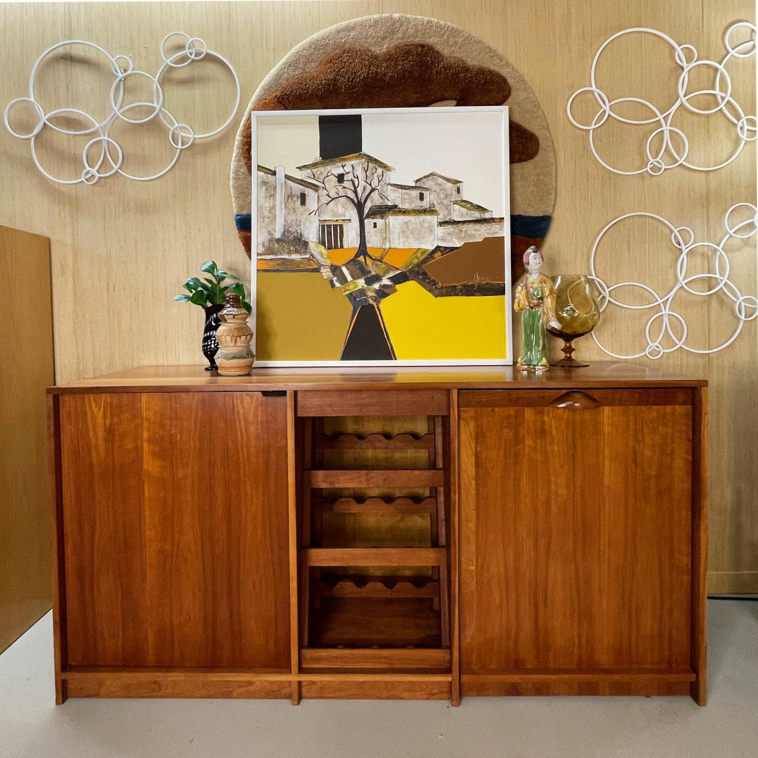 The painting is a mid-century modern piece created by artist Frank Walcutt in the 1970s. The artwork features a homestead scene with houses, trees, and a serene landscape, showcasing elements of nature. The color palette includes white, brown, and