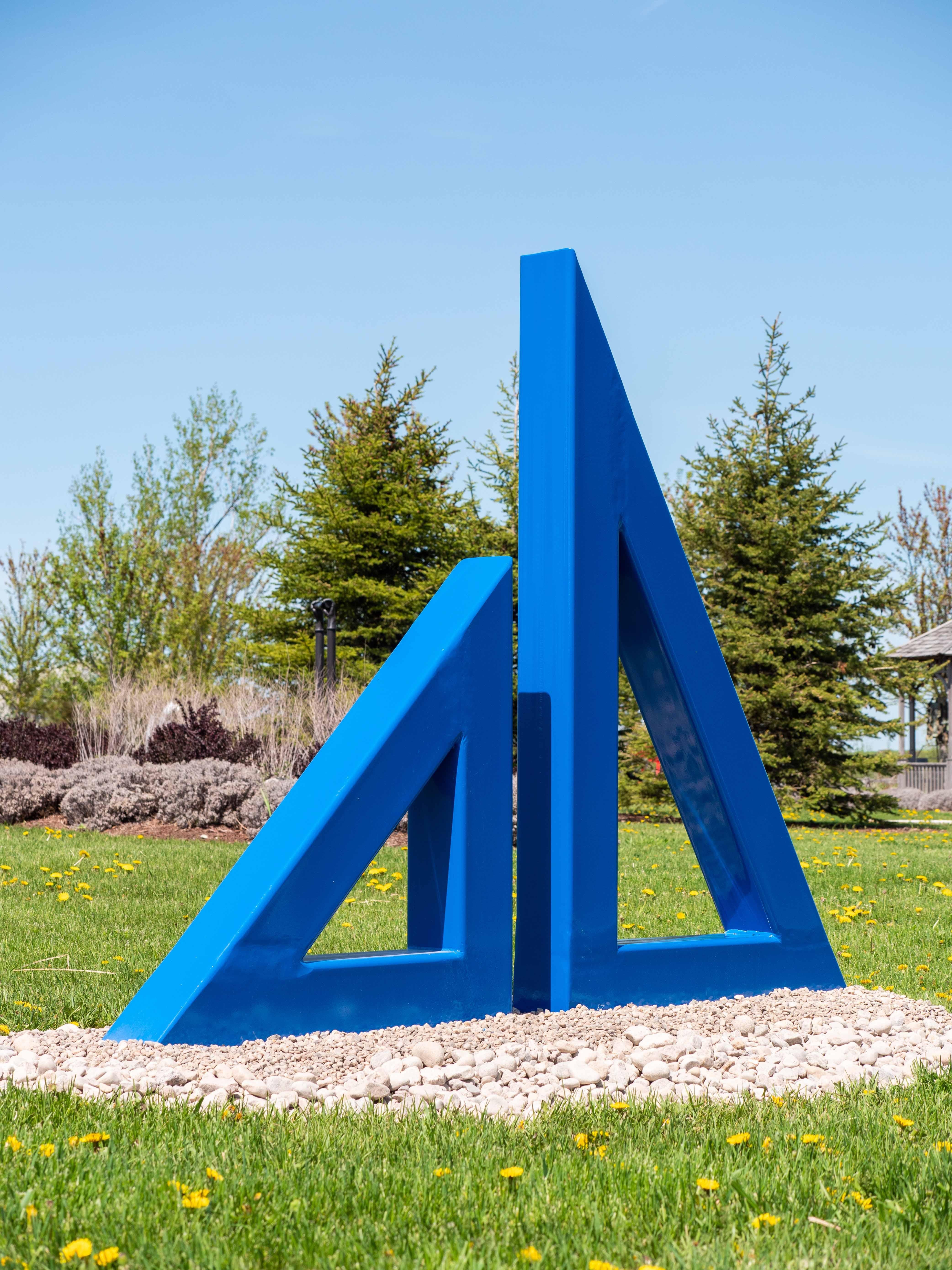 Zwei auffällige, königsblau lackierte Stahldreiecke in unterschiedlichen Größen, deren Mittelpunkte ausgeschnittene Dreiecke sind, stehen nebeneinander in dieser neuen Außenskulptur des kanadischen Künstlers Fraser Radford. Radford arbeitet in