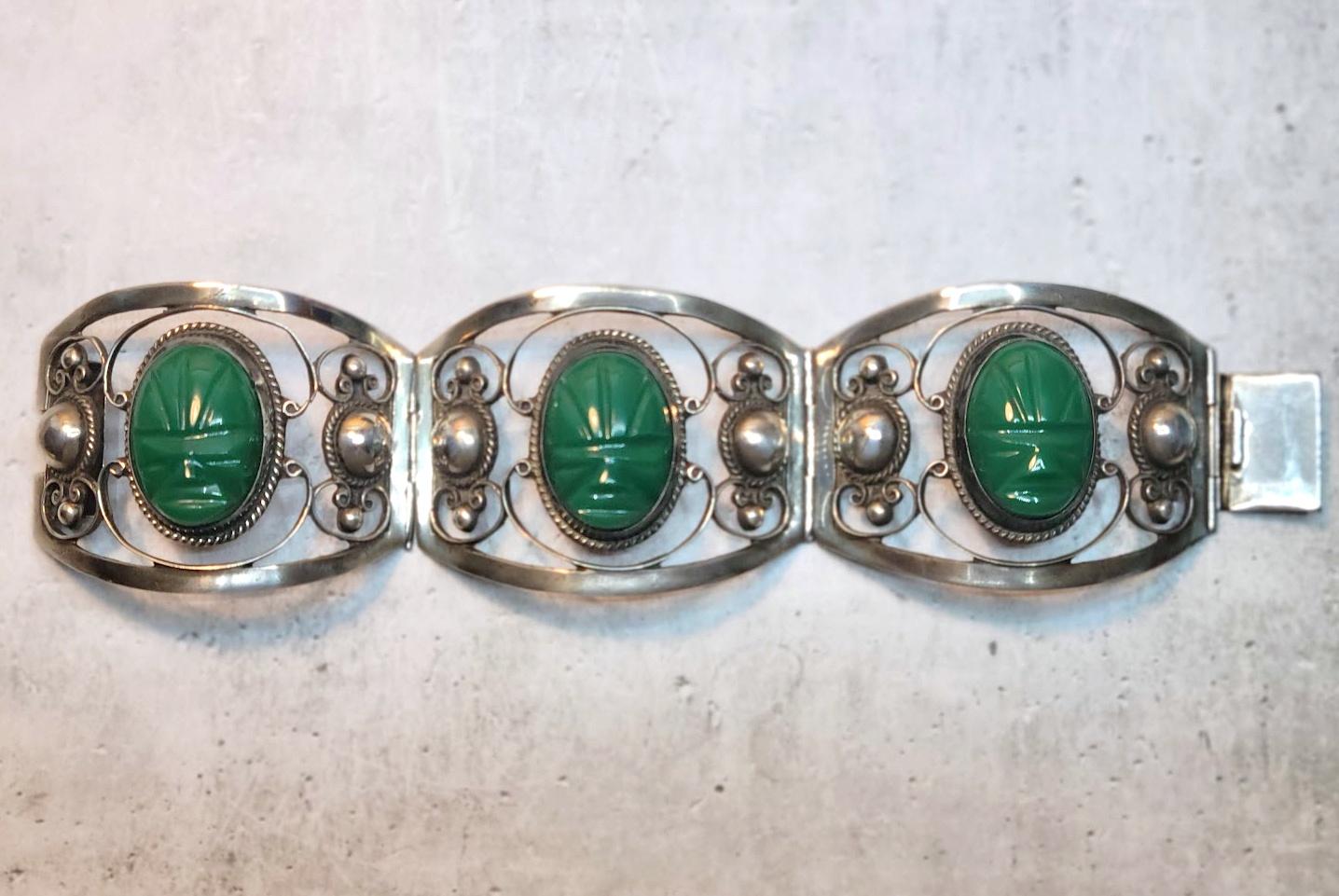 A Mexican bracelet comprised of fine sterling silver with green onyx cabochons in the form of Aztec masks. The bracelet has decorative silver panels with heavy beading, swirls, and oval onyx Aztec masks.

This vintage panel bracelet was made in