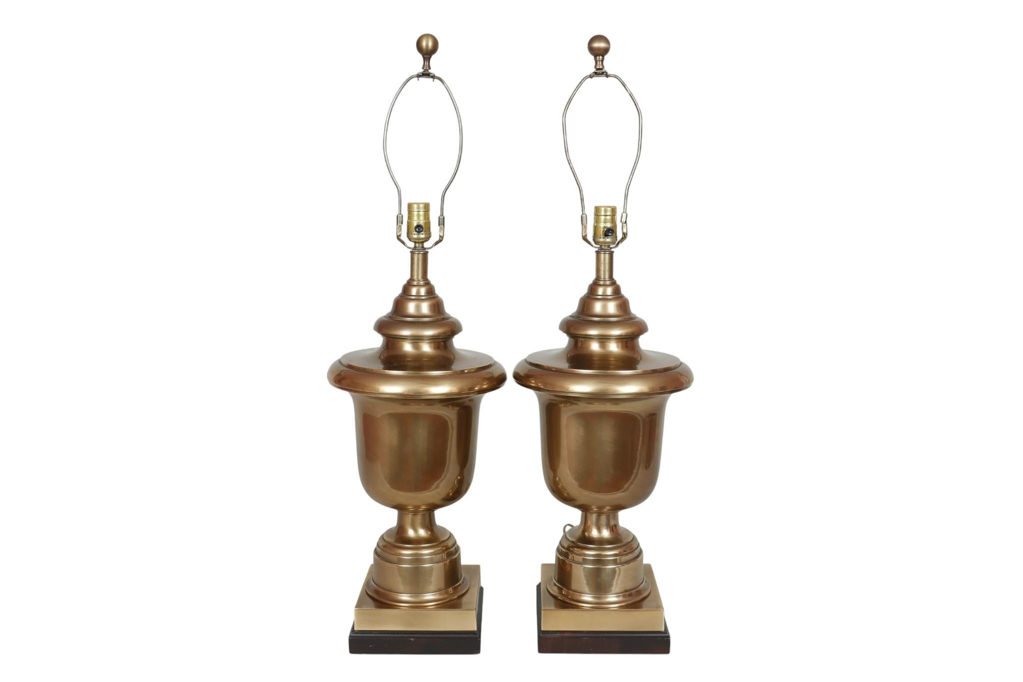 Paire de lampes de table en forme d'urne en laiton, fabriquées par Frederick Cooper. De grandes urnes en laiton tourné sont posées sur des bases carrées biseautées. Les abat-jours en forme de cloche en crème claire sont bordés d'un galon tressé et