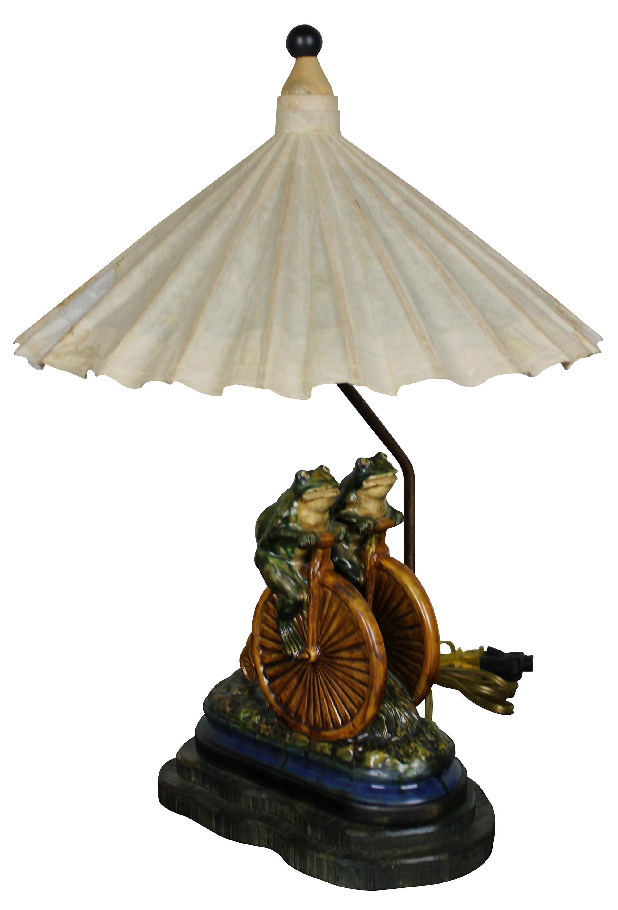 Late 20th century Frederick cooper desk lamp. A figural design in ceramic feautiring two frogs riding a penny farthing bicycle over wood cut base. Includes two way lights and a parasol shade. Measure: 23