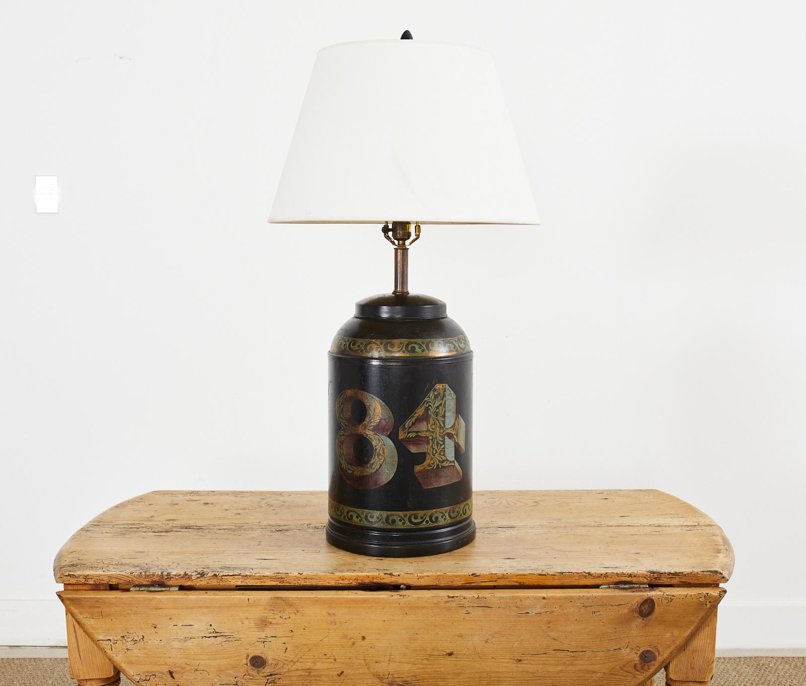 Vintage tea canister tole lamp finished overall in black with a decorative gilt number 84 and foliate designs in gold, green, and oxblood color. Brass hardware with harp and finial. Lamp shade not included.