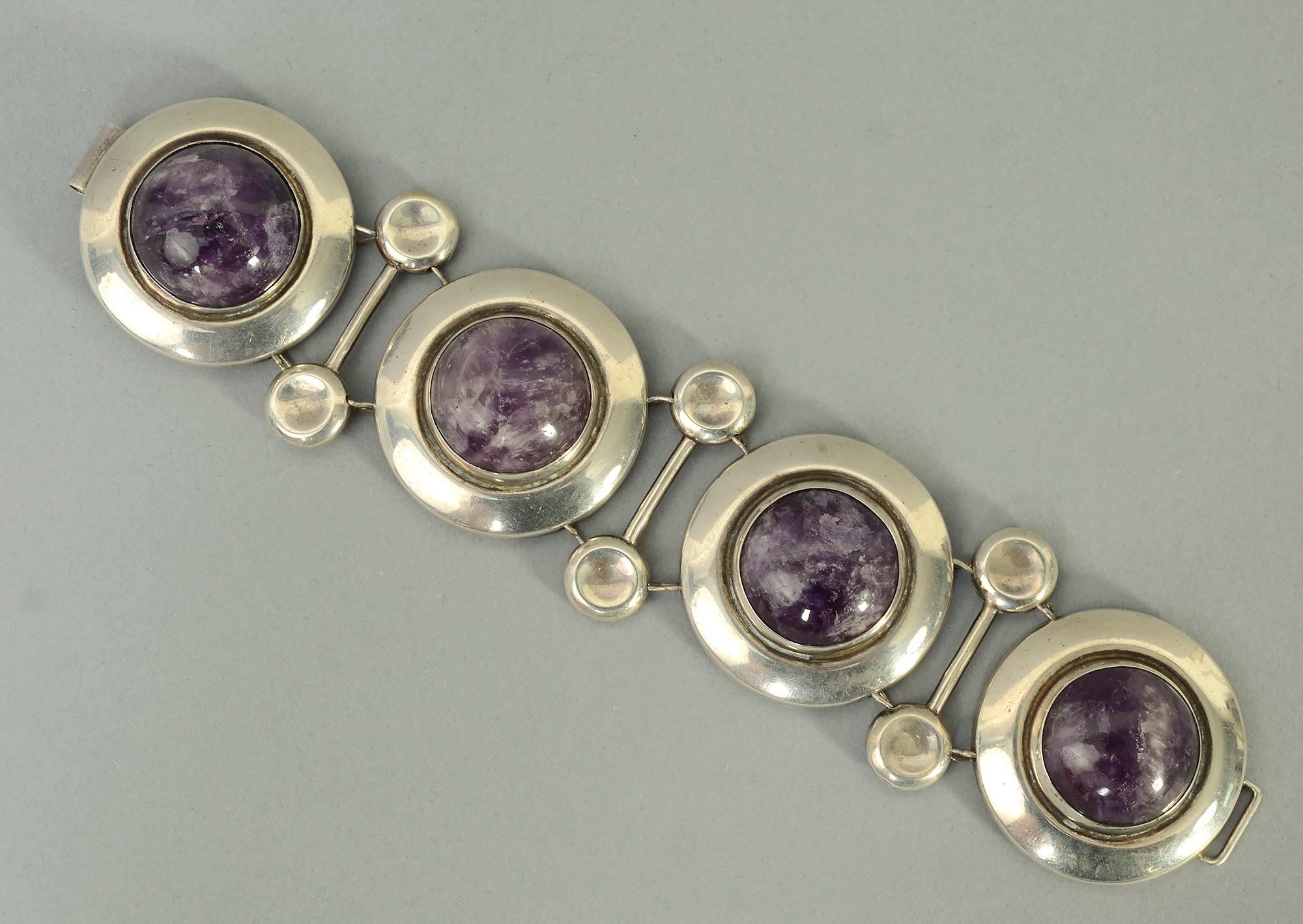 This dramatic bracelet by Frederick Davis (1880 - 1961) is as unusual as it is beautiful.
The bracelet consists of four silver discs in which are set cabochon amethysts. The links are separated by silver bars, the ends of which repeat the circle