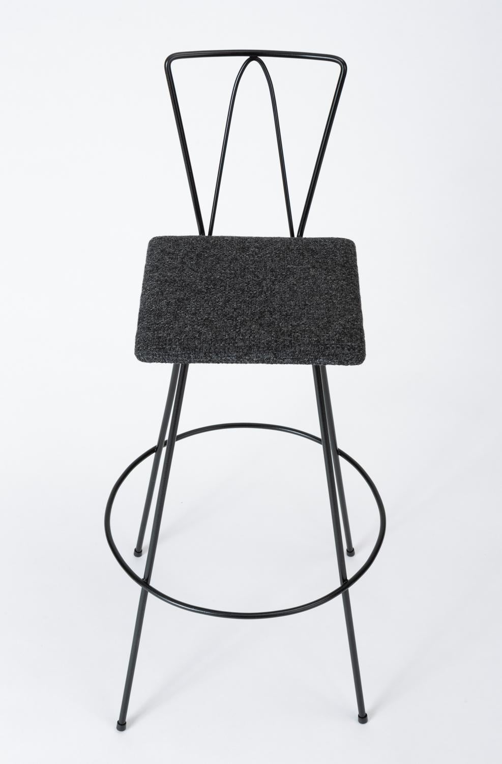 A modernist bar stool with a slender profile in bent, powder-coated metal. The stool has four angled legs, encircled by a support bar and footrest. The feet are capped with rubber tips to protect surfaces. The square seat is upholstered in a dark