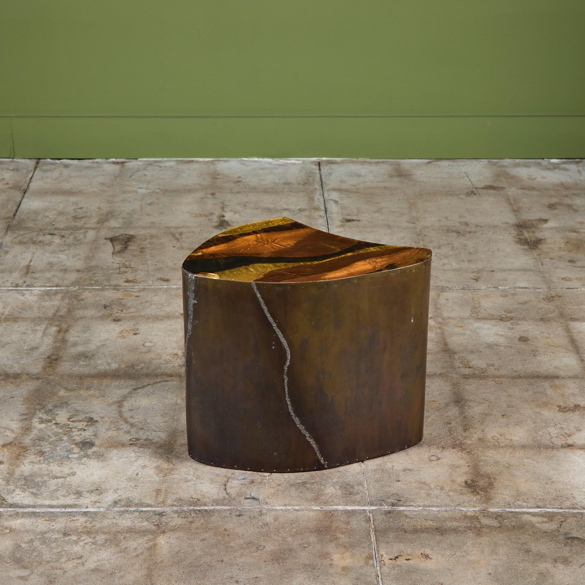 Mesa auxiliar de forma libre revestida de latón con tablero de metal mixto. La mesa tiene un detalle de remaches de latón a lo largo de los lados. El tablero de la mesa presenta láminas de latón y cobre con una capa de resina. Hay cuatro