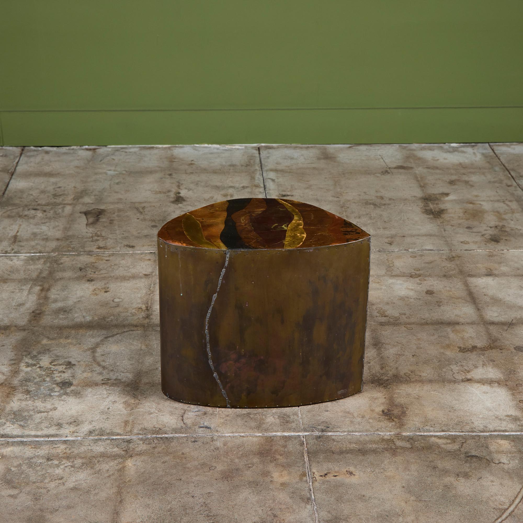 Mesa auxiliar de forma libre revestida de latón con tablero de metal mixto. La mesa tiene un detalle de remaches de latón a lo largo de los lados. El tablero de la mesa presenta láminas de latón y cobre con una capa de resina. Hay cuatro