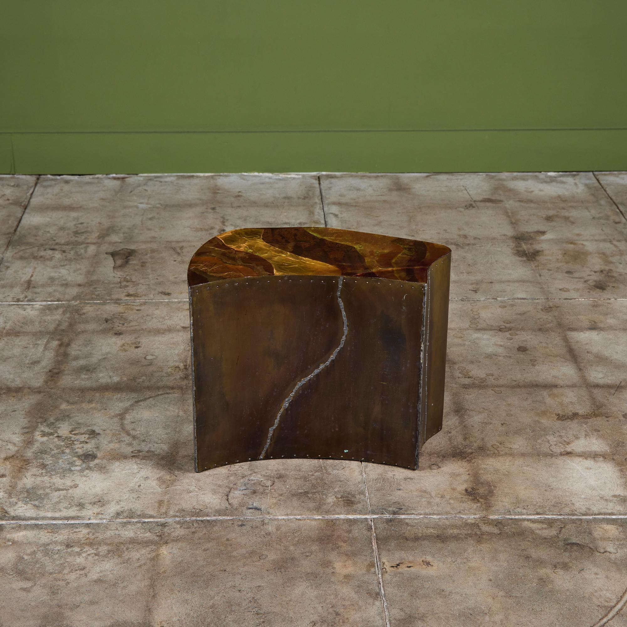Mesa auxiliar de forma libre revestida de latón con tablero de metal mixto. La mesa tiene un detalle de remaches de latón a lo largo de los lados. El tablero de la mesa presenta láminas de latón y cobre con una capa de resina. Hay cuatro