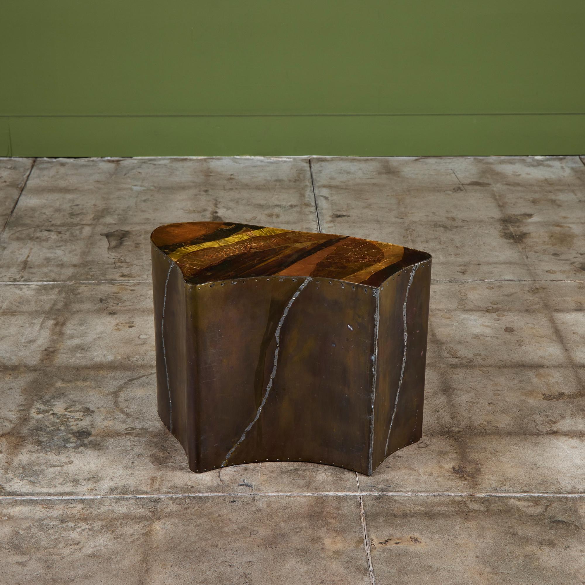 Mesa auxiliar de forma libre revestida de latón con tablero de metal mixto. La mesa tiene un detalle de remaches de latón a lo largo de los lados. El tablero de la mesa presenta láminas de latón y cobre con una capa de resina. Hay cuatro