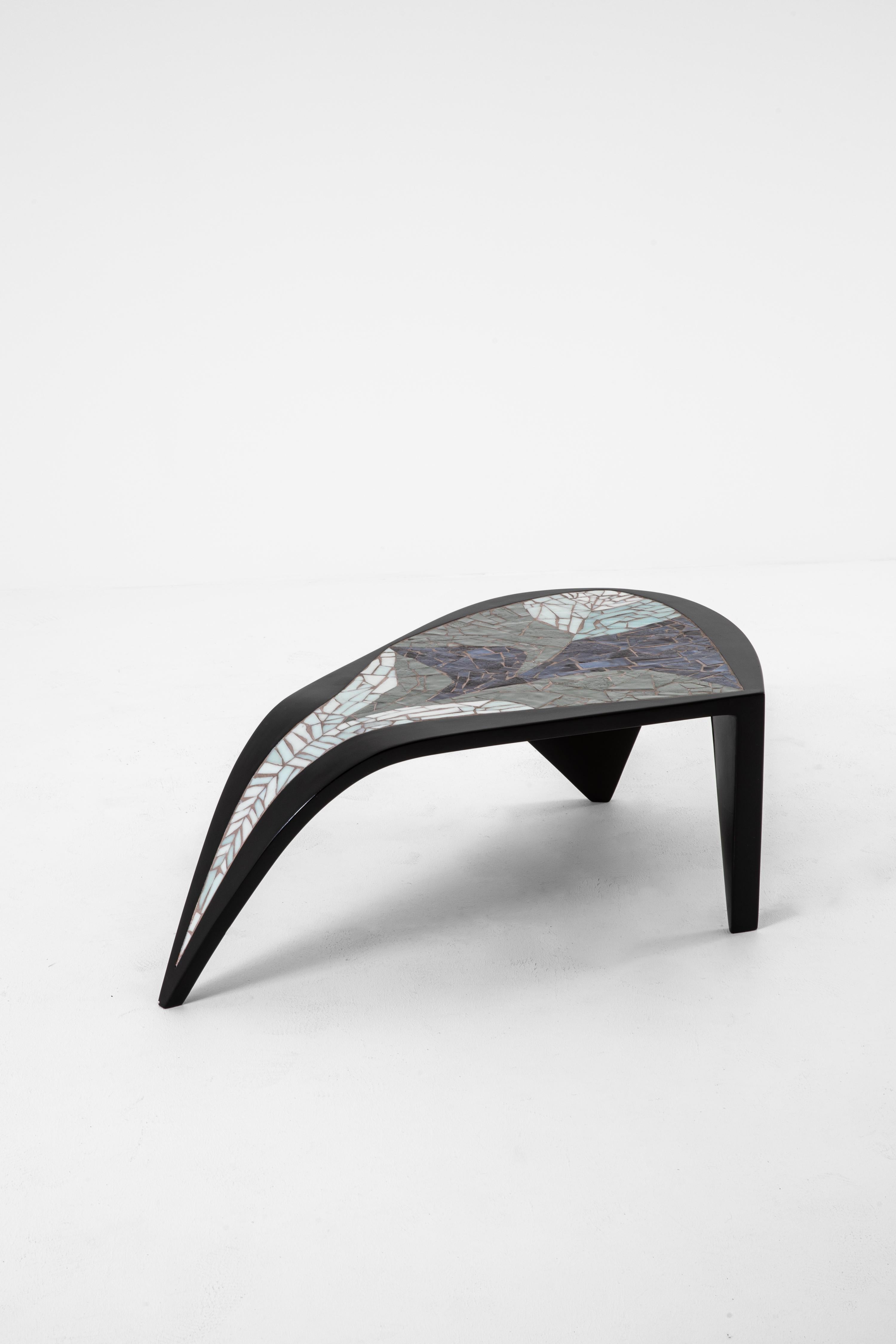 The tripod coffee table, created in 1986, embodies the free and organic forms that define Valentine Schlegel's work. Supported by three legs of lacquered wood, it unfolds into a softly contoured top, adorned with a mosaic of Venetian glass whose