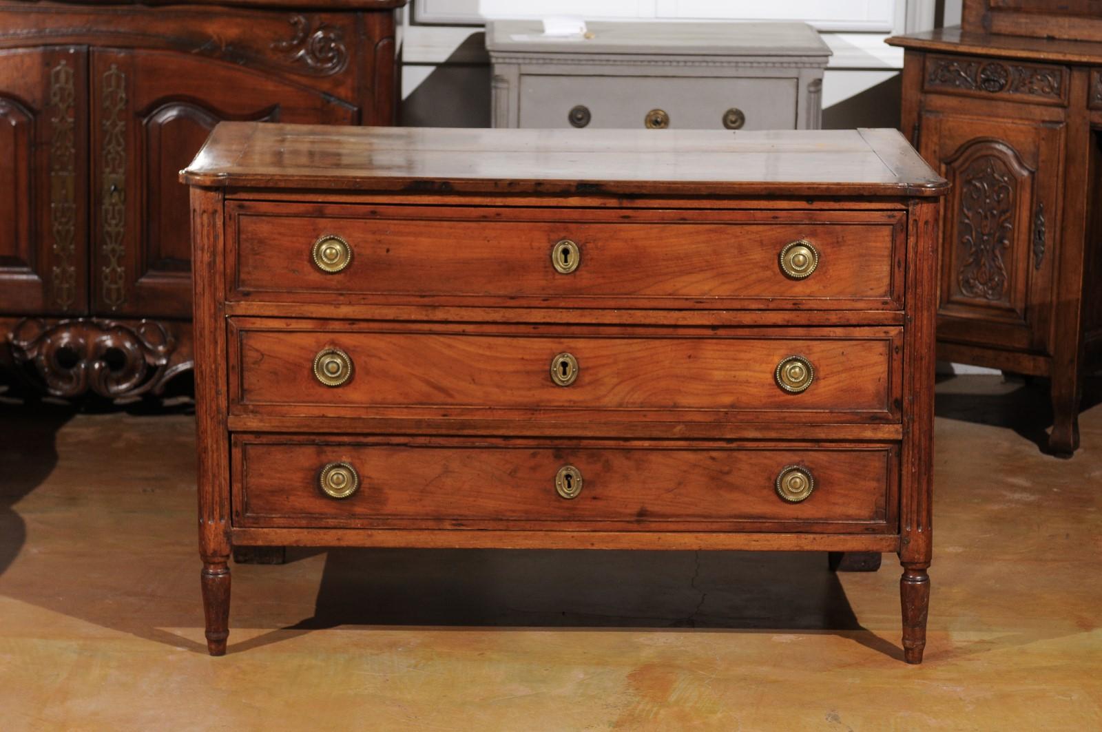 A French Louis XVI period walnut commode from the 18th century, with three drawers and fluted accents. Born in France during the early years of the reign of King Louis XVI, this walnut commode features a rectangular planked top with rounded corners