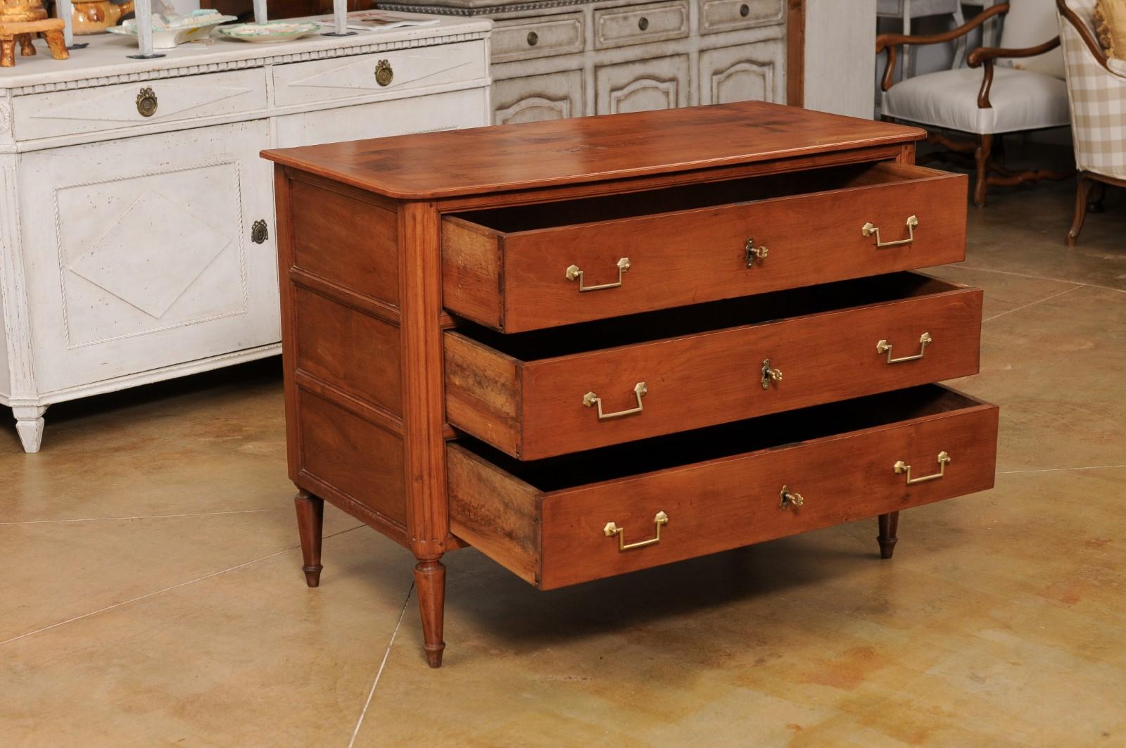 A French Louis XVI period cherry commode from the late 18th century, with three drawers and fluted side posts. Created in France during the last decade of the 18th century, this cherry commode features a rectangular top with rounded corners in the