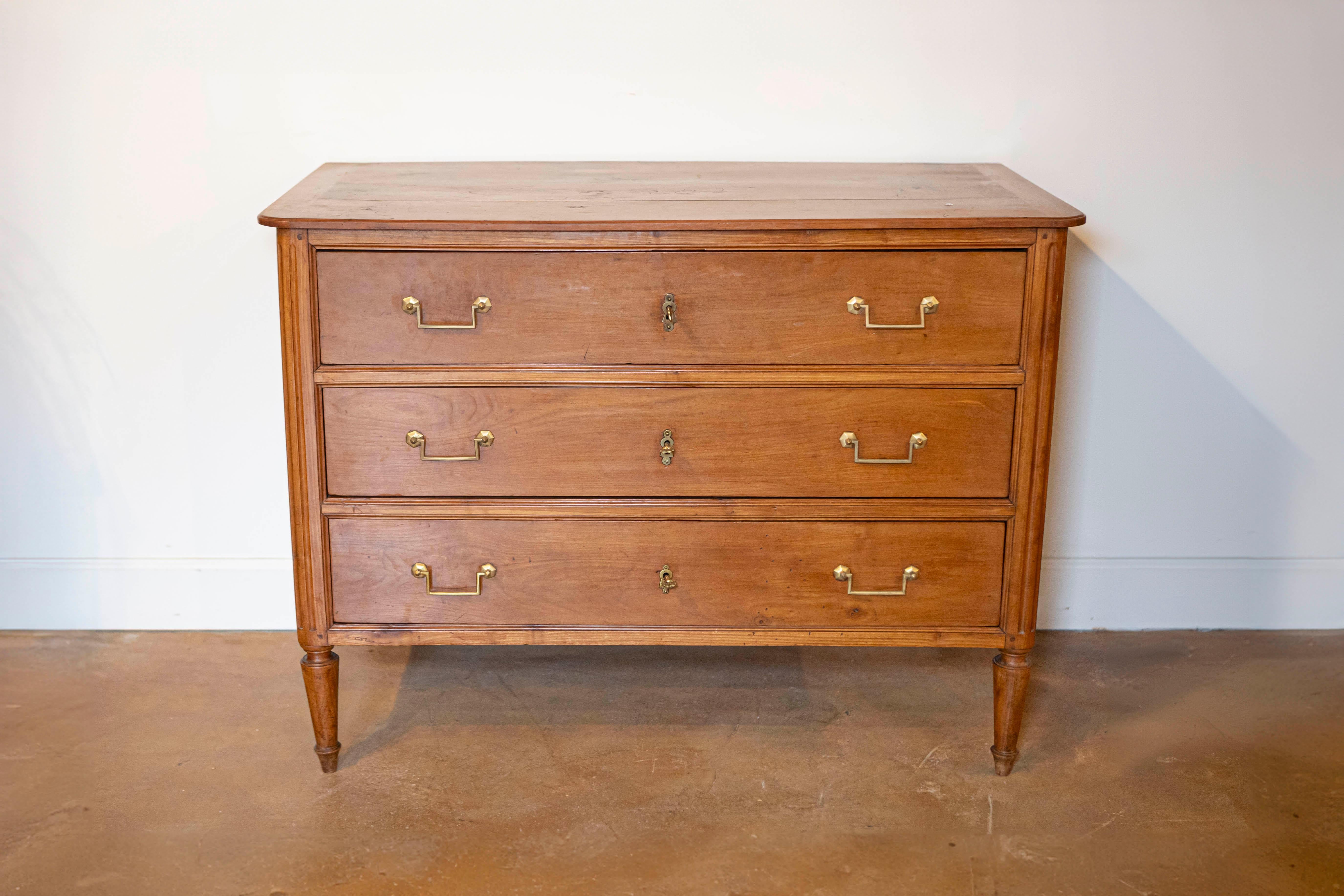 A French Louis XVI period cherry commode from the late 18th century, with three drawers and fluted side posts. Created in France during the last decade of the 18th century, this cherry commode features a rectangular top with rounded corners in the