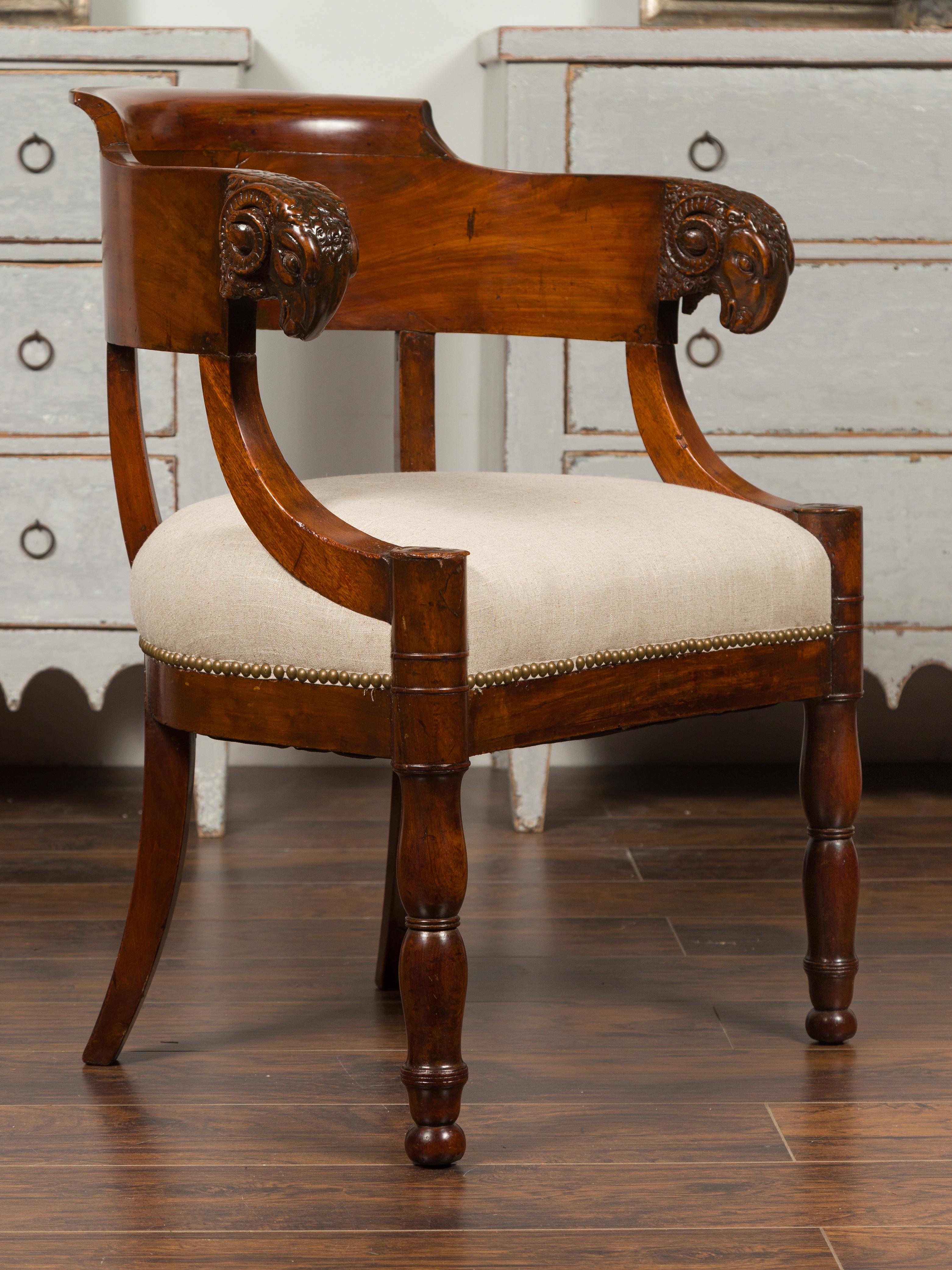A French Restauration mahogany armchair from the 19th century, with carved rams' heads and new upholstery. Born in France during the Restauration period, this mahogany armchair features a wraparound back adorned with exquisitely carved rams' heads.
