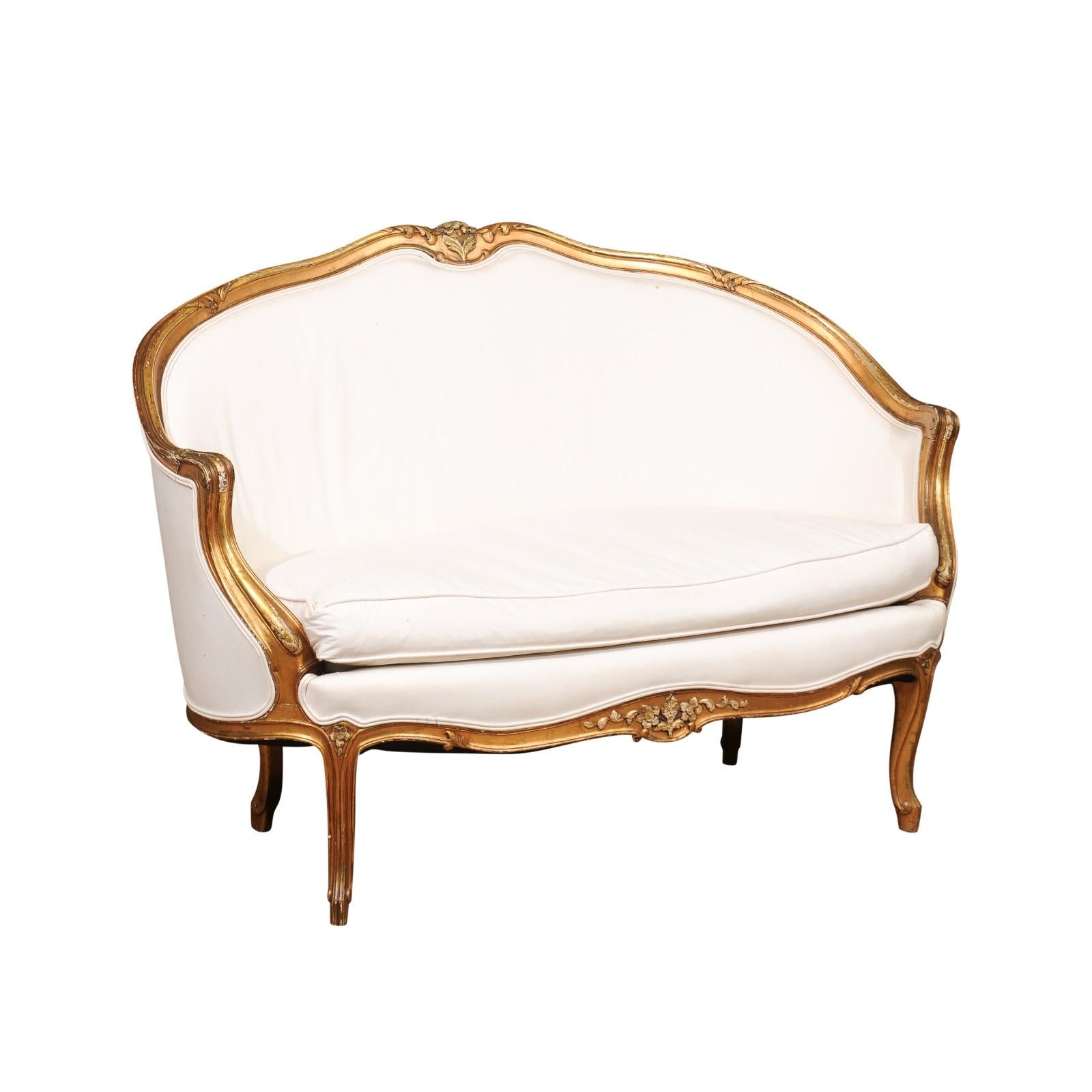 A French Louis XV style giltwood petite upholstered sofa from the mid-19th century. This small size French canapé features a giltwood frame showing the typical curvy lines of the Louis XV era. The crest is carved with a delicate floral motif