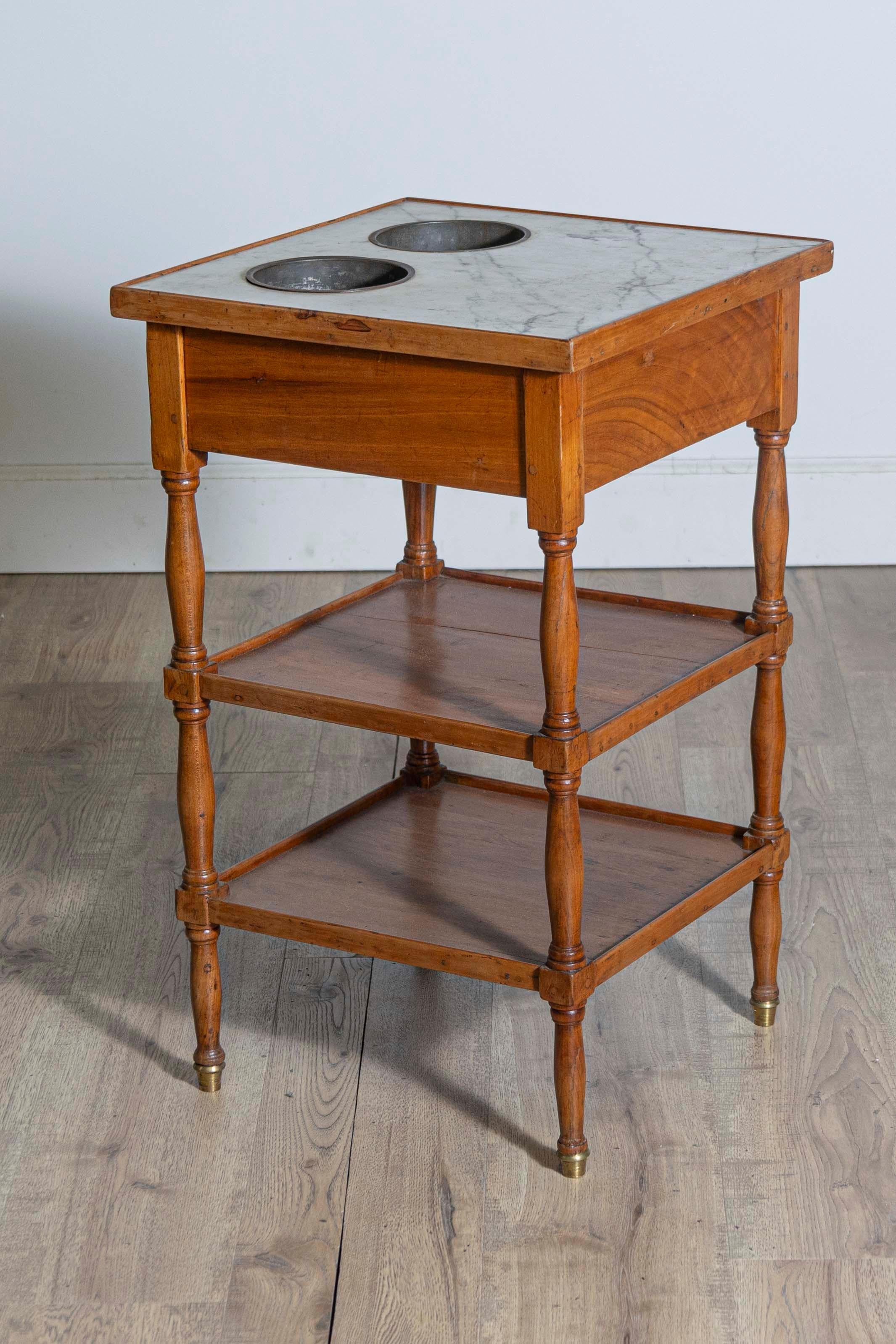 A French cherry tiered rafraichissoire wine table circa 1890 with two inset wine buckets, white veined marble top and single drawer. Immerse yourself in the antique charm of French craftsmanship with this cherry tiered rafraichissoire wine table,