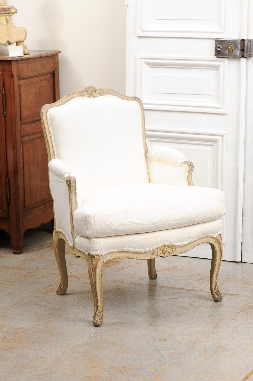 A French Louis XV style painted and carved wooden bergère chair from the late 19th century, with lace fabric and distressed patina. Created in France during the last quarter of the 19th century, this painted bergère features a slightly slanted back