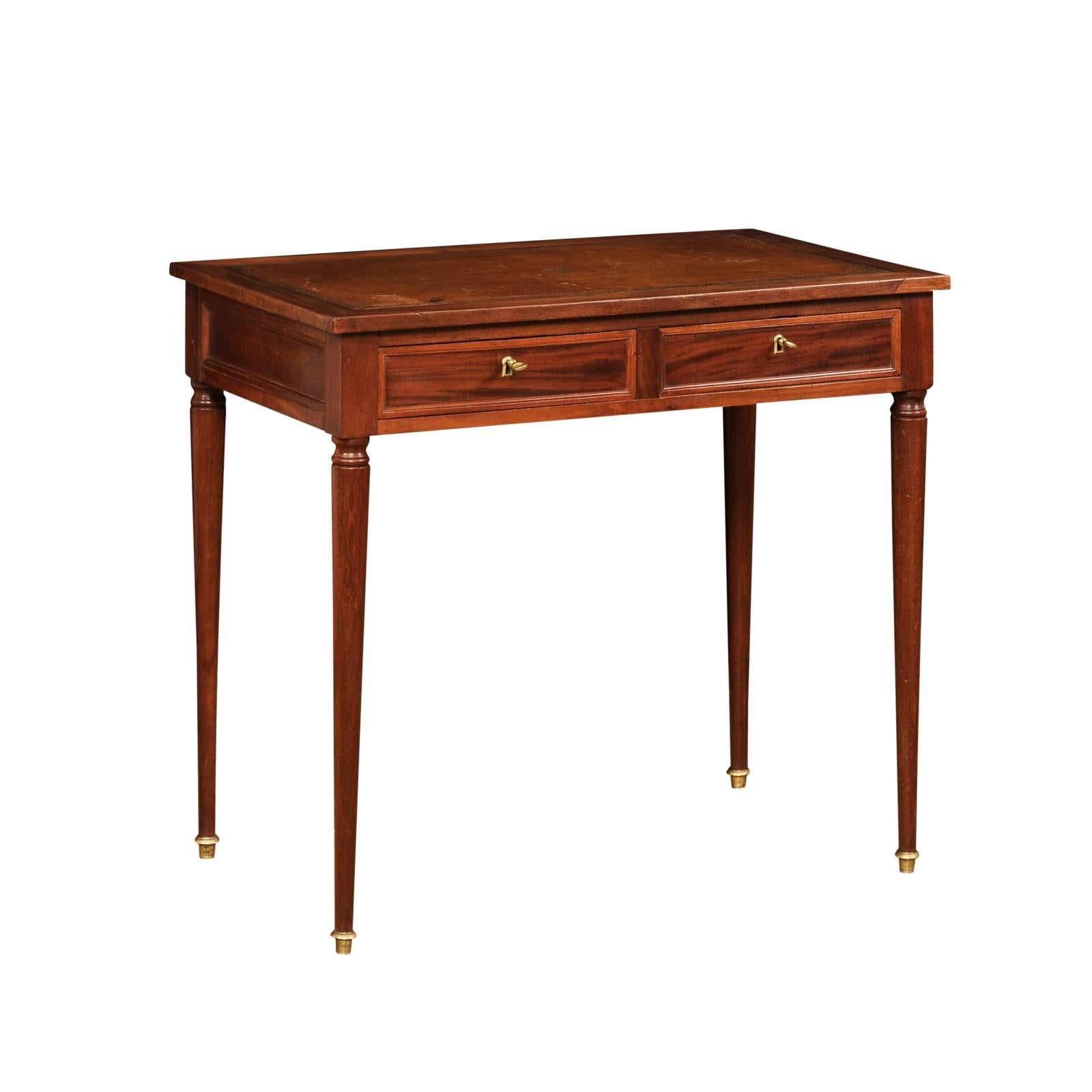 A petite French Louis XVI style mahogany and mahogany veneer desk from the late 19th century, with brown leather top, two drawers and cylindrical legs on bronze feet. Created in France during the last quarter of the 19th century, this petite Louis