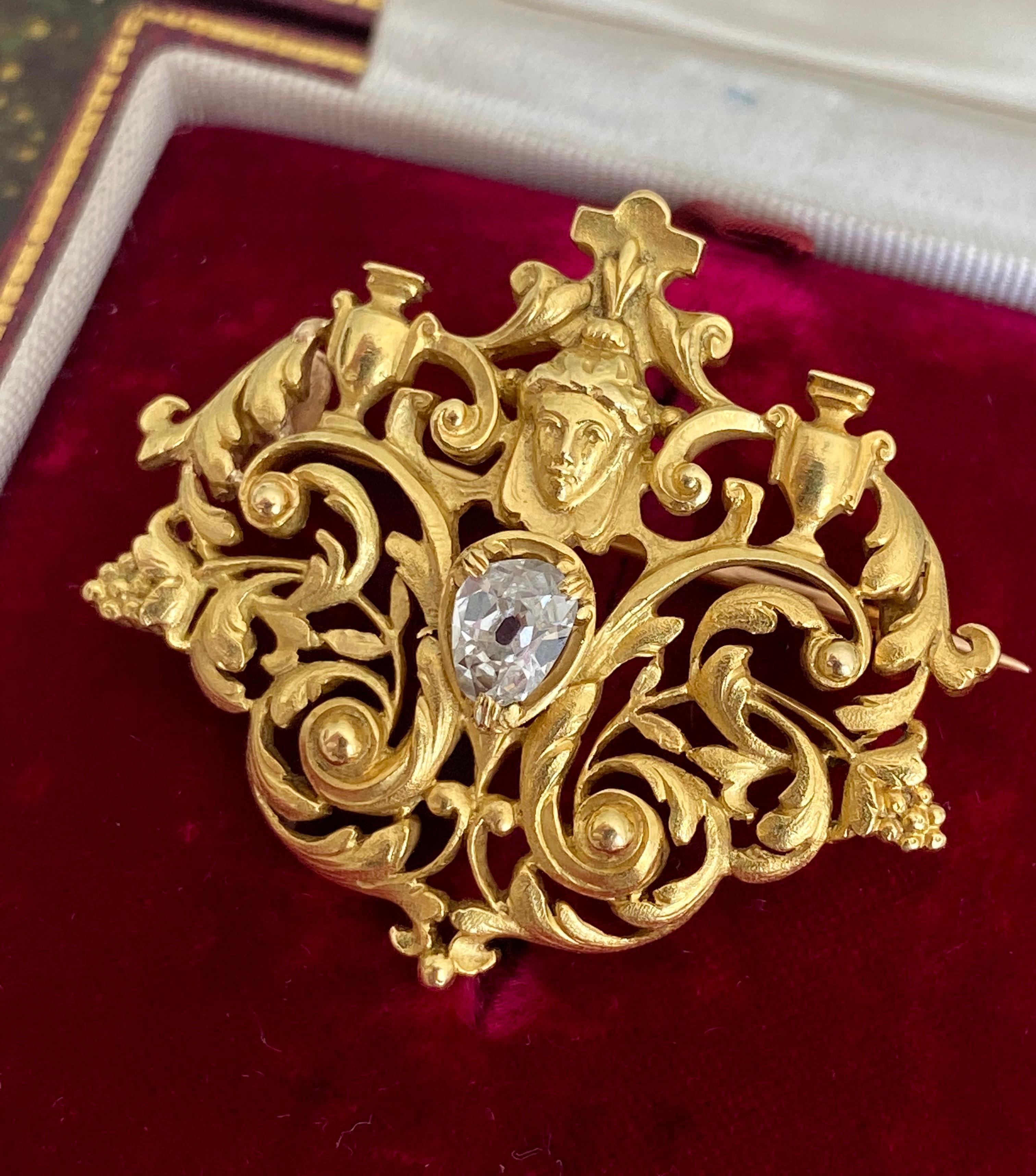 This exceptional French brooch centers on a serene beauty with upswept hair perched above an old pear cut diamond. Symmetrically arranged openwork foliate flow around her, further embellished by a tiny clusters of grapes and a pair of handled urns.