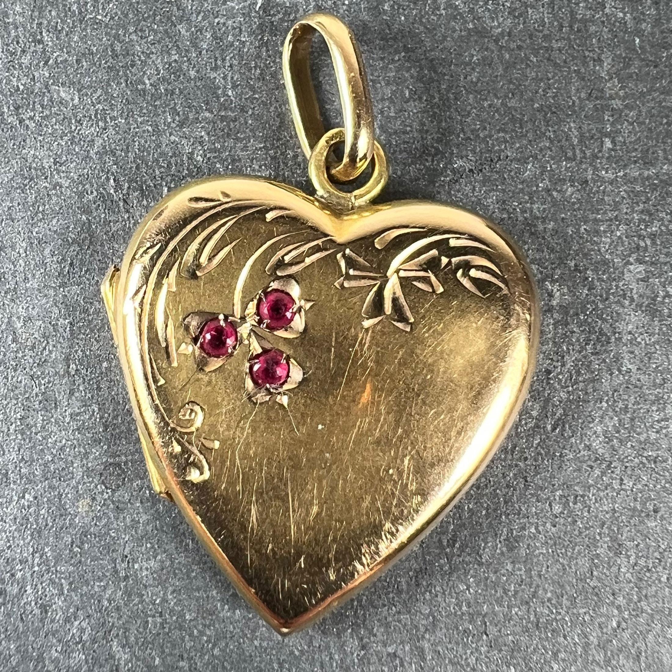 A French 18 karat (18K) yellow gold charm pendant designed as a heart-shaped locket with engraved floral and lucky three-leaf clover decoration, set with three red rubies to a trefoil design. Stamped with the eagle's head for French manufacture and