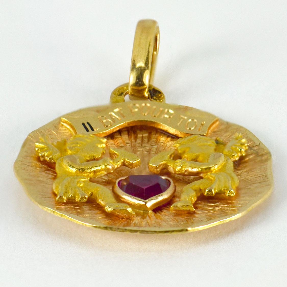 A French 18 karat (18K) yellow gold charm pendant designed as a disk with two yellow gold cupid figures with hammers beating a synthetic ruby love heart. Above them a enamelled scroll reads 'Il bat pour toi', the whole meaning 'My heart beats for