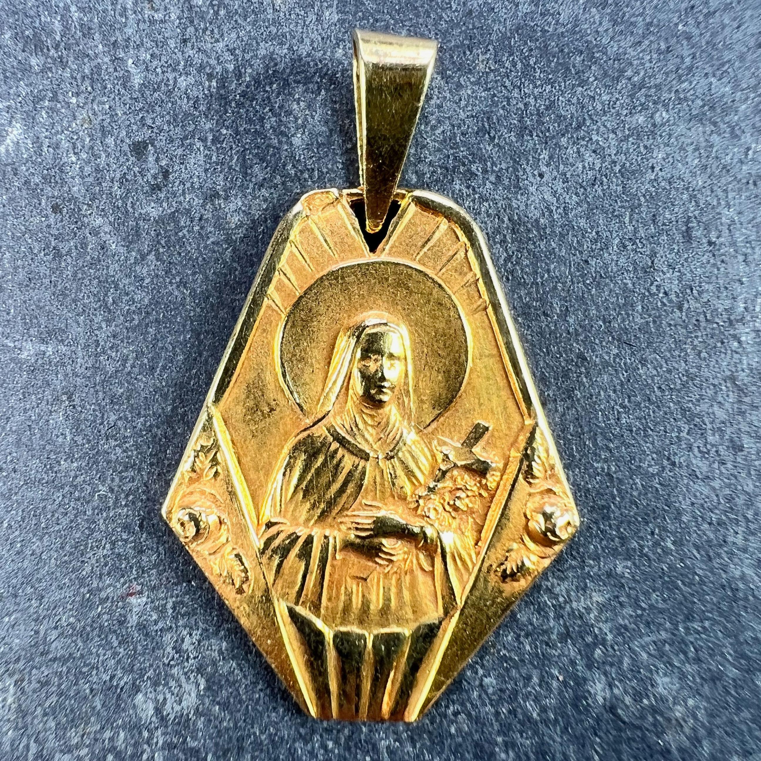 A French 18 karat (18K) yellow gold charm pendant designed as an hexagonal medal depicting Saint Therese with a halo holding a crucifix and some roses, the surround with architectural detail and more roses. Stamped with the eagle's head for 18 karat